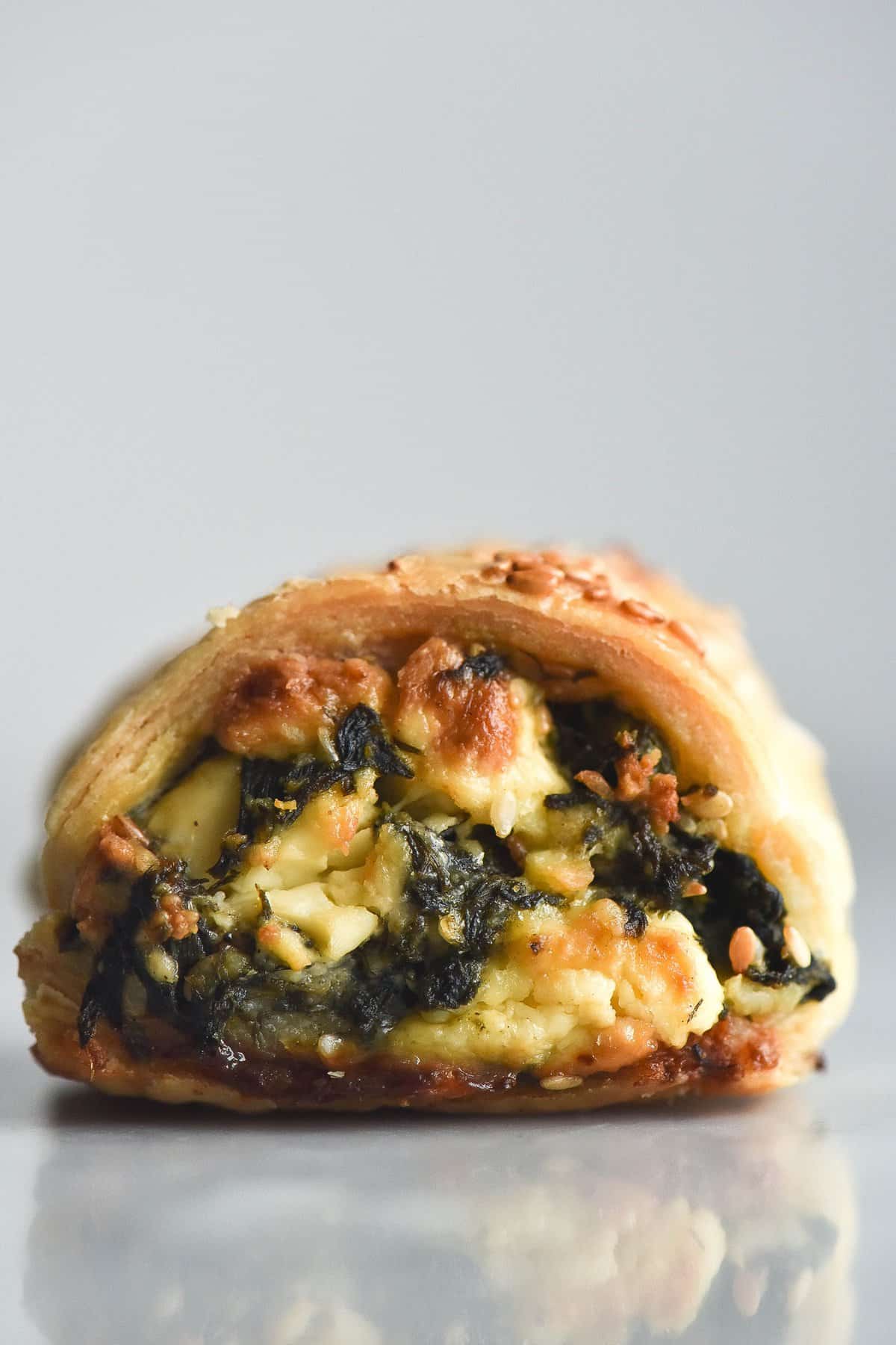 A side on macro photo of a gluten free spinach and feta sausage roll. The roll sits atop a white marble table against a white background. Golden feta and pine nuts poke out of the filling, and the top of the pastry is sprinkled with toasted sesame seeds