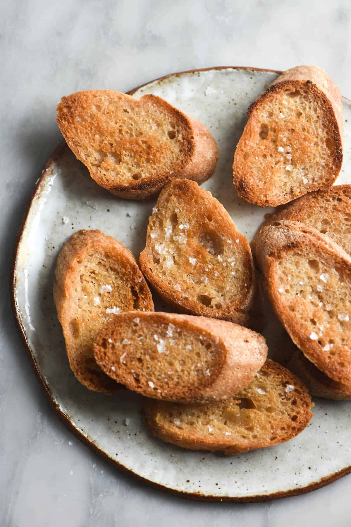 An aerial view of a plate of FODMAP friendly, gluten free garlic ghee infused garlic bread. The bread is covered in flaky salt and sits atop a small white ceramic plate on a white marble table