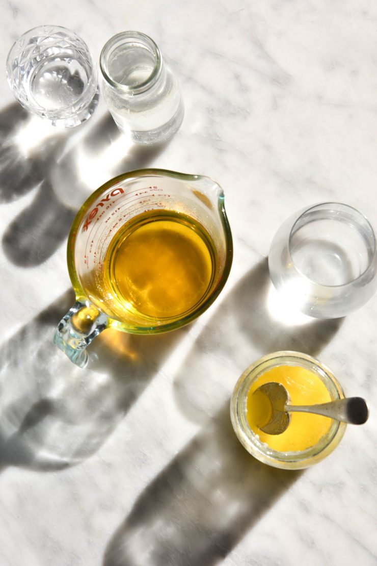 An aerial view of two jugs of freshly made garlic infused ghee sit on a white marble table in harsh sunlight. They are surrounded by water glasses and the light shines through creating shadows on the marble