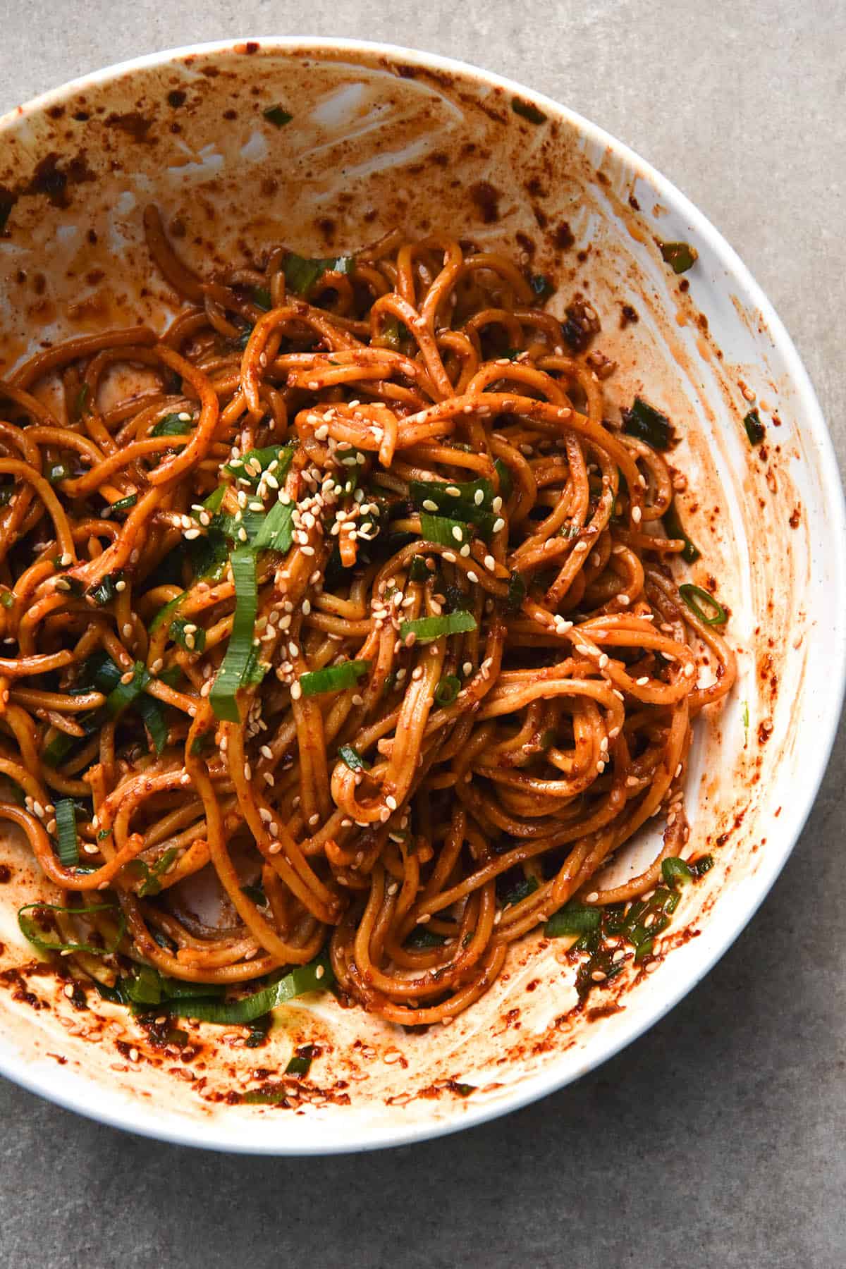 A close up, aerial view of a messy plate of FODMAP friendly chilli oil noodles. The noodles are casually strewn in the white bowl and their red hue contrasts with the spring onion greens and sesame seeds that garnish them. The bowl sits atop a light grey slate tile.