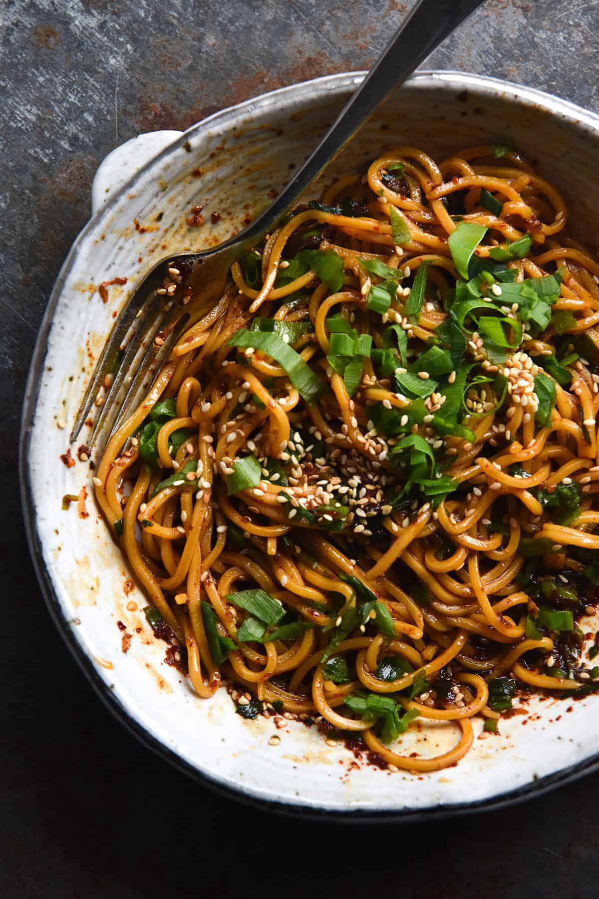 A close up aerial view of FODMAP friendly chilli oil noodles in a white ceramic bowl. A fork pokes out of the top left hand side of the bowl, extending across to the right. The bowl sits atop a dark blue metal backdrop. The noodles are casually strewn in the bowl and topped with sesame seeds and spring onion greens
