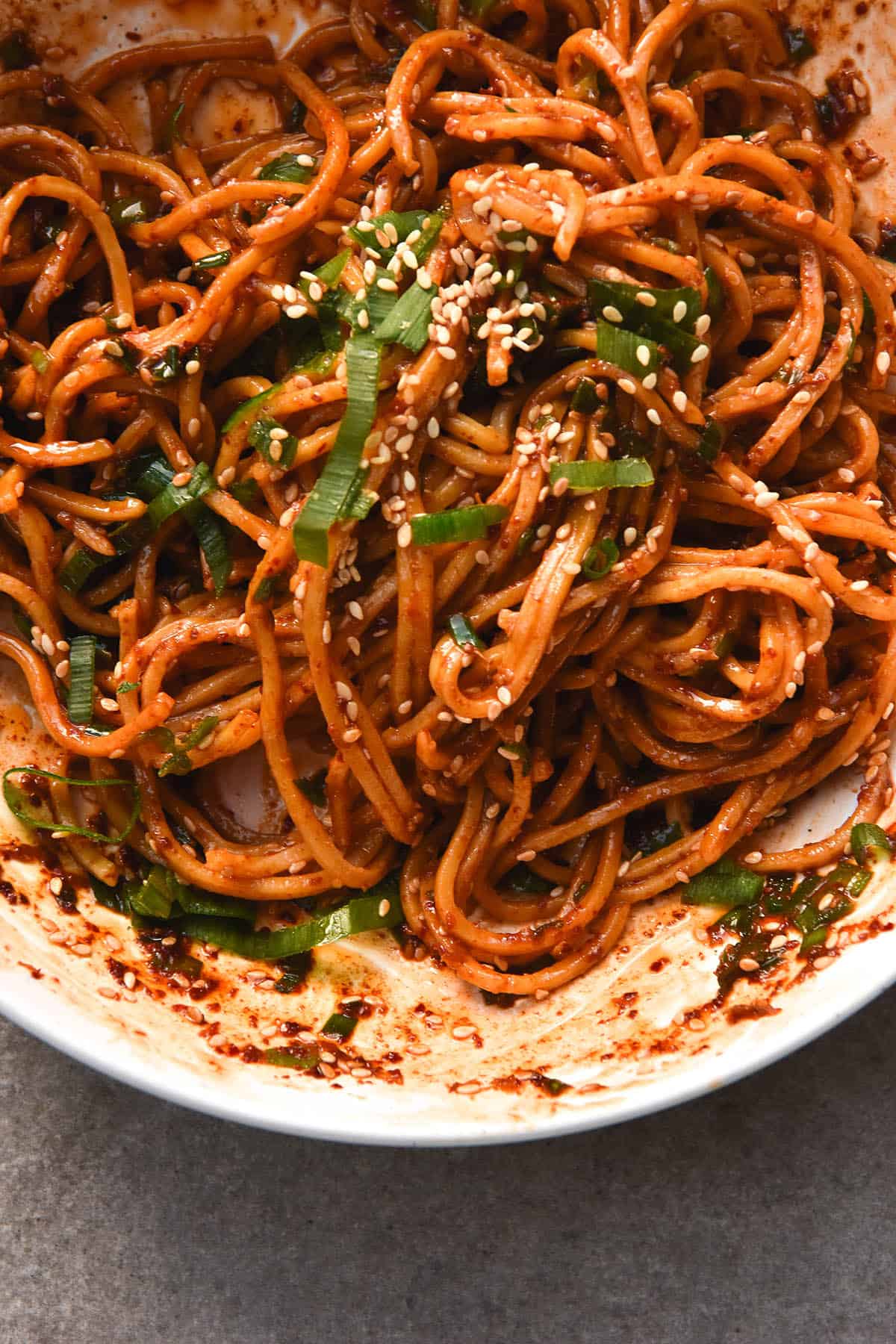 A close up aerial view of a messy white bowl of FODMAP friendly garlic oil noodles. The noodles are a deep red hue and topped with sesame seeds and spring onion greens. The grey stone backdrop is visible underneath the bowl in the bottom of the image