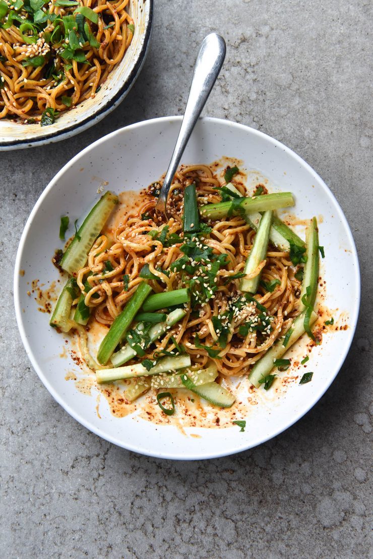 An aerial view of two white bowls of FODMAP-friendly chilli oil noodles. The bowls sit atop a light grey stone background, and one bowl is the centre of the image, while the other peeks out of the top lefthand corner. The bowl in the centre has a fork in it, poking out the top right hand side of the image. The noodles are casually topped with spring onion greens, toasted sesame seeds and cucumber batons