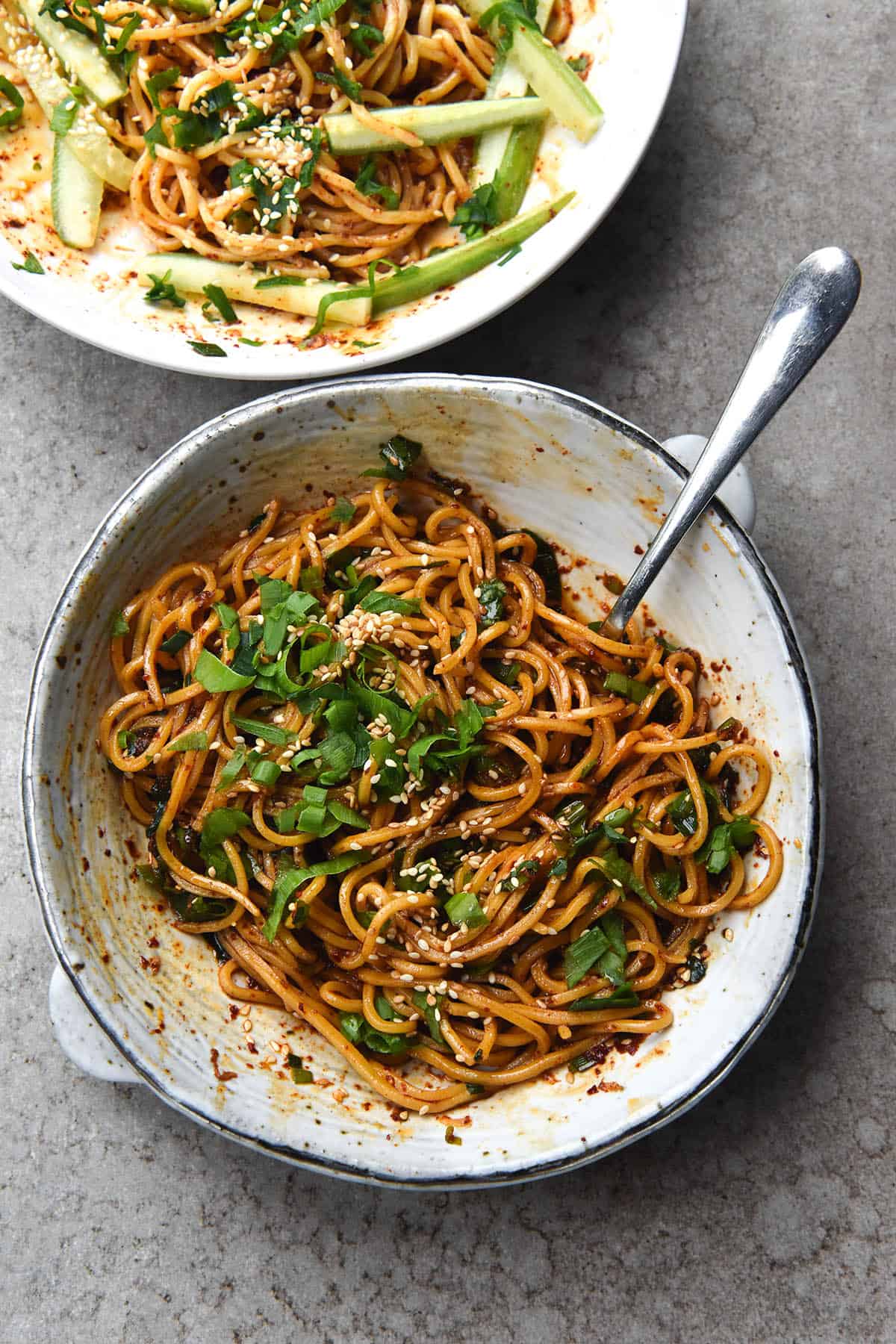 An aerial view of two white bowls of FODMAP-friendly chilli oil noodles. The bowls sit atop a light grey stone background, and one bowl is the centre of the image, while the other peeks out of the top lefthand corner. The bowl in the centre has a fork in it, poking out the top right hand side of the image. The noodles are casually topped with spring onion greens and toasted sesame seeds