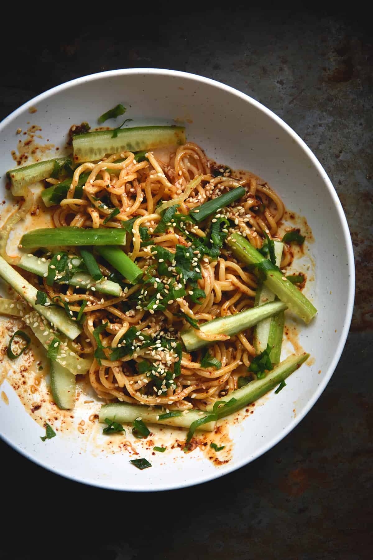 An aerial view of a white bowl of FODMAP friendly chilli oil noodles. The noodles are casually strewn about in the bowl, and topped with cucumber batons, spring onion greens and sesame seeds. The bowl sits atop a dark grey steel backdrop, which contrasts with the white bowl and vibrant chilli red tinted hue of the noodles