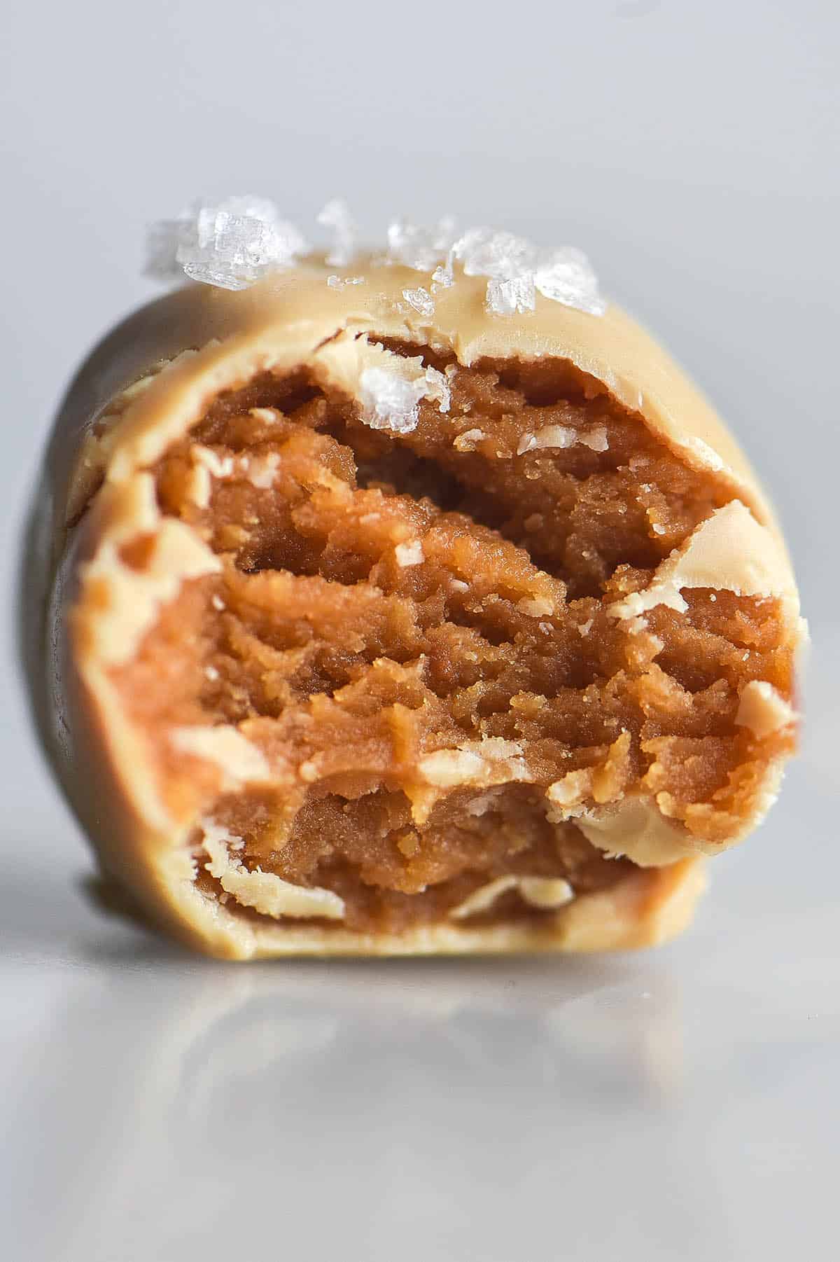 A close up side on image of a peanut butter truffle coated in white chocolate. A bite has been taken, revealing the peanut butter centre, and sea salt flakes adorn the top. The truffle sits atop a white marble table against a white backdrop