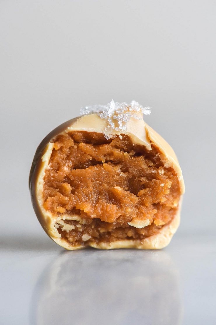 A close up side on image of a peanut butter truffle coated in white chocolate. A bite has been taken, revealing the peanut butter centre, and sea salt flakes adorn the top. The truffle sits atop a white marble table against a white backdrop