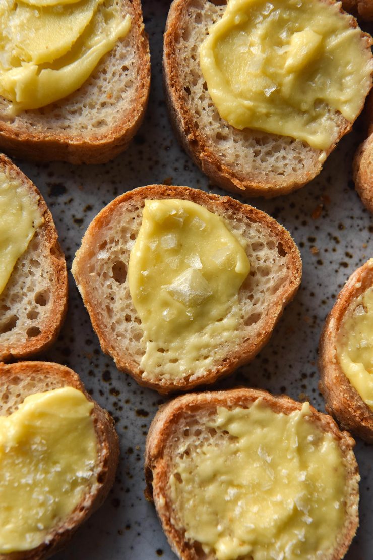 An aerial view of a plate of sliced baguette rounds slathered in whipped garlic infused ghee and topped with flaky sea salt. The baguette rounds sit atop a white speckled plate which is visible between the pieces of baguette