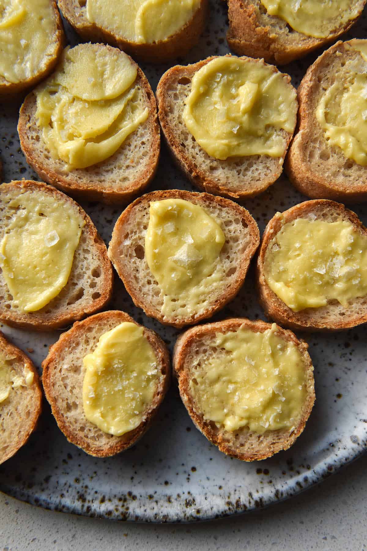 An aerial view of a plate of sliced baguette rounds slathered in whipped garlic infused ghee and topped with flaky sea salt. The baguette rounds sit atop a white speckled plate which is visible at the bottom of the image. The plate sits on a white countertop.