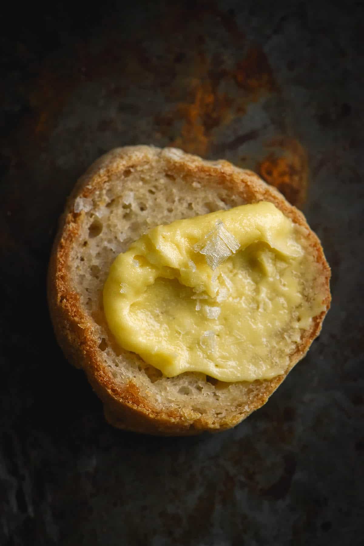 An aerial close up view of a round slice of baguette topped with a swirl of whipped garlic infused ghee and flaky sea salt. The baguette round sits atop a dark grey backdrop with rusty splotches