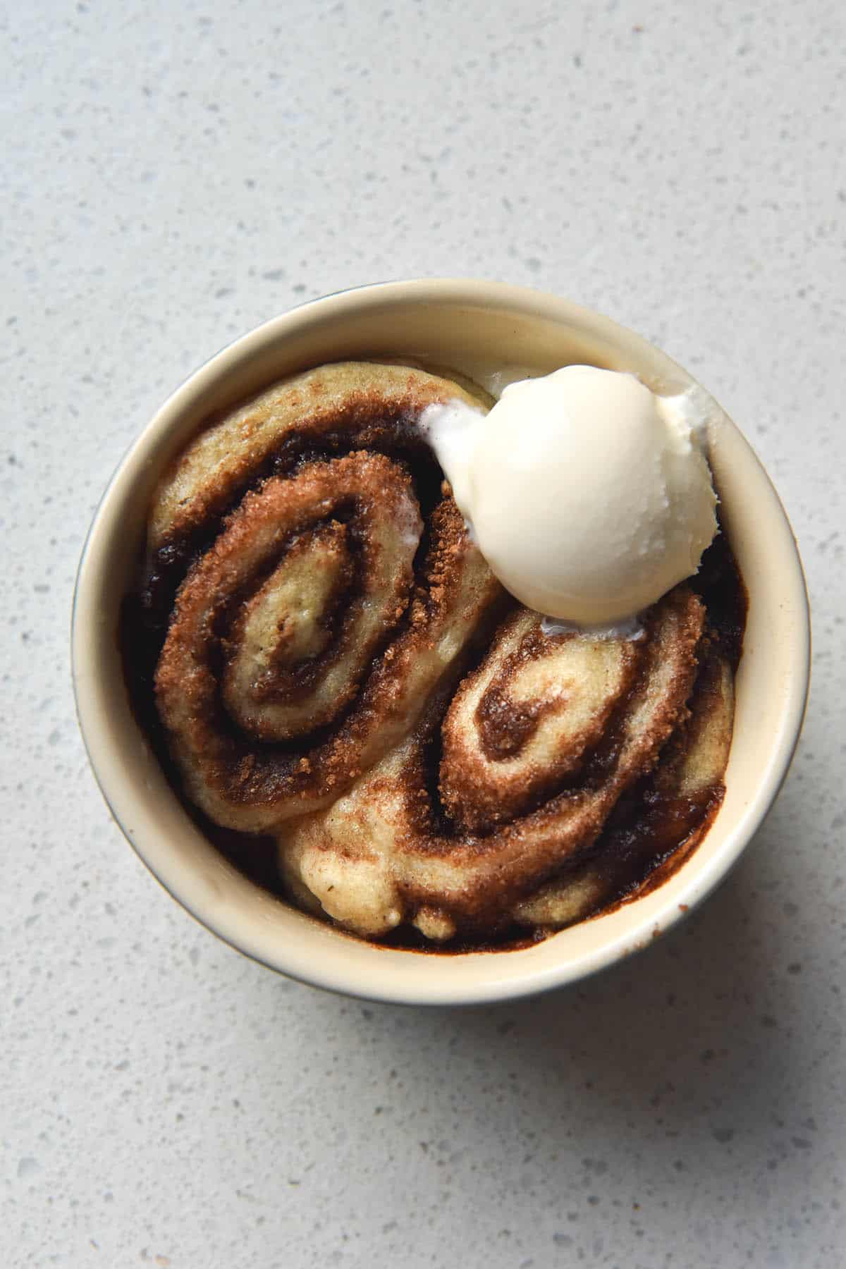 An aerial close up view of a gluten-free vegan microwave cinnamon scroll. The scroll has been cooked in a beige ramekin, which sits atop a white speckled ceramic plate. There are two scrolls snuggled into the dish and are topped with a small scoop of vanilla ice cream