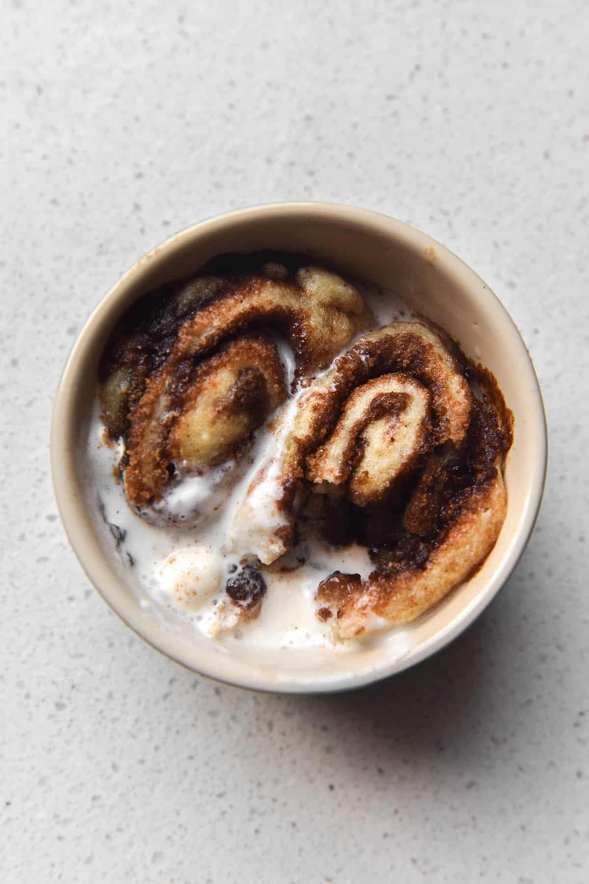 An aerial close up view of a gluten-free vegan microwave cinnamon scroll. The scroll has been cooked in a beige ramekin, which sits atop a white kitchen counter. There are two scrolls snuggled into the dish and are topped with a small scoop of melted vanilla ice cream