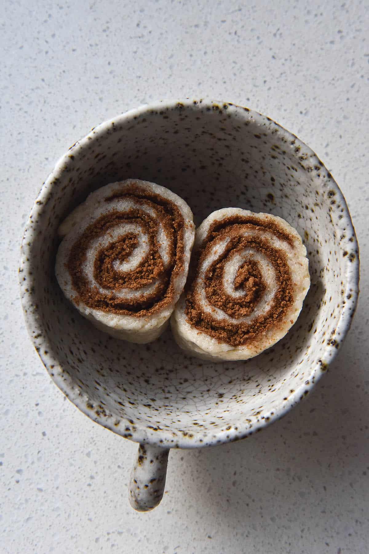 An aerial image of an uncooked gluten free, vegan microwave cinnamon scroll. The scroll has been sliced in half, and the two parts sit in a white ceramic speckled mug, cinnamon scroll side up. They are full to the brim with cinnamon sugar. The mug sits atop a white kitchen bench