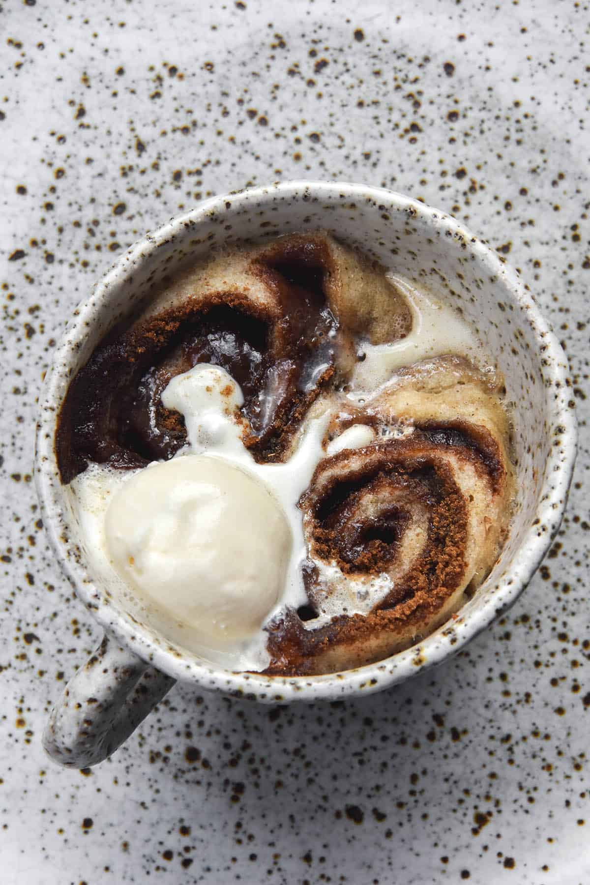 An aerial image of a vegan, gluten free microwave cinnamon scroll in a white speckled ceramic mug. The scroll is oozing cinnamon sugar and topped with melting vanilla ice cream. It sits atop a white speckled ceramic plate