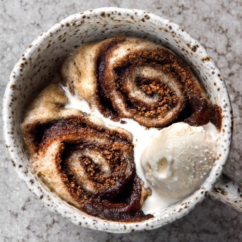 An aerial image of a vegan, gluten free microwave cinnamon scroll in a white speckled ceramic mug. The scroll is oozing cinnamon sugar and topped with melting vanilla ice cream. It sits atop a light grey stone backdrop