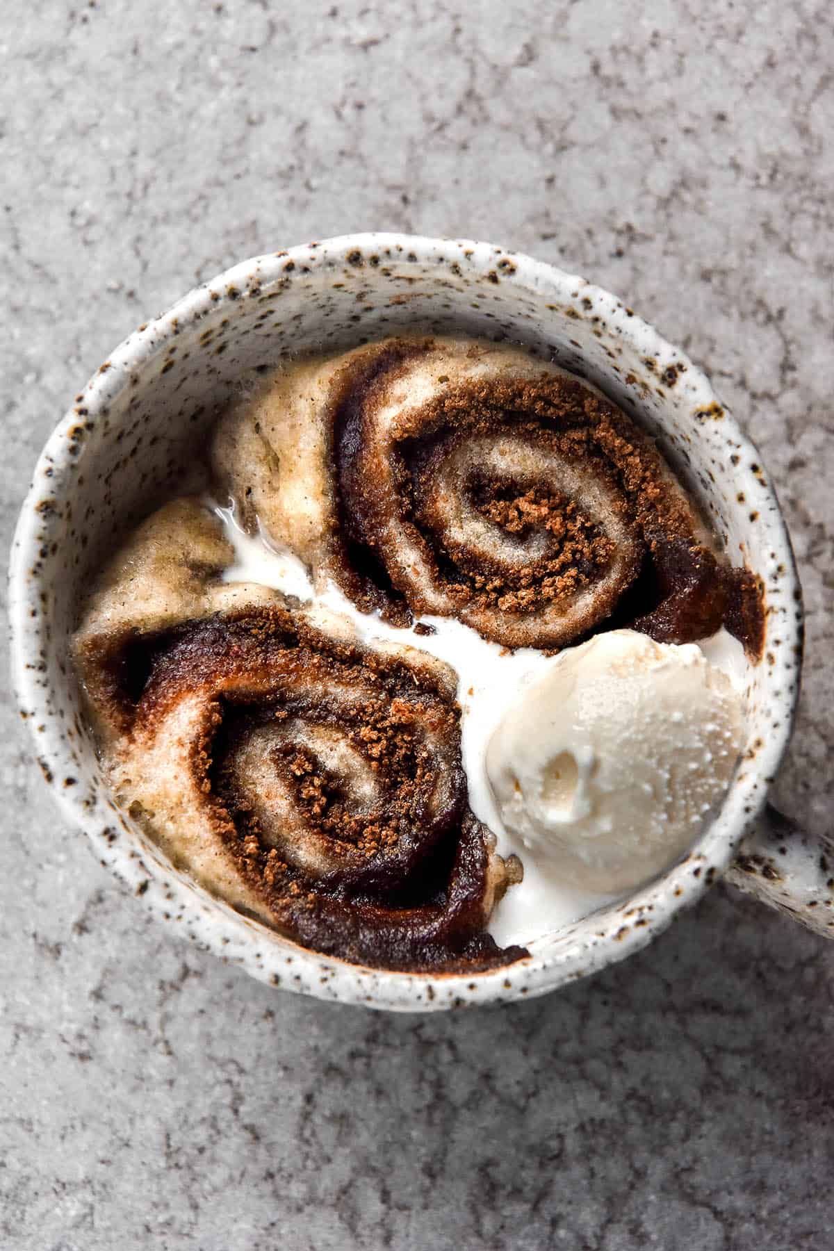 An aerial image of a vegan, gluten free microwave cinnamon scroll in a white speckled ceramic mug. The scroll is oozing cinnamon sugar and topped with melting vanilla ice cream. It sits atop a light grey stone backdrop