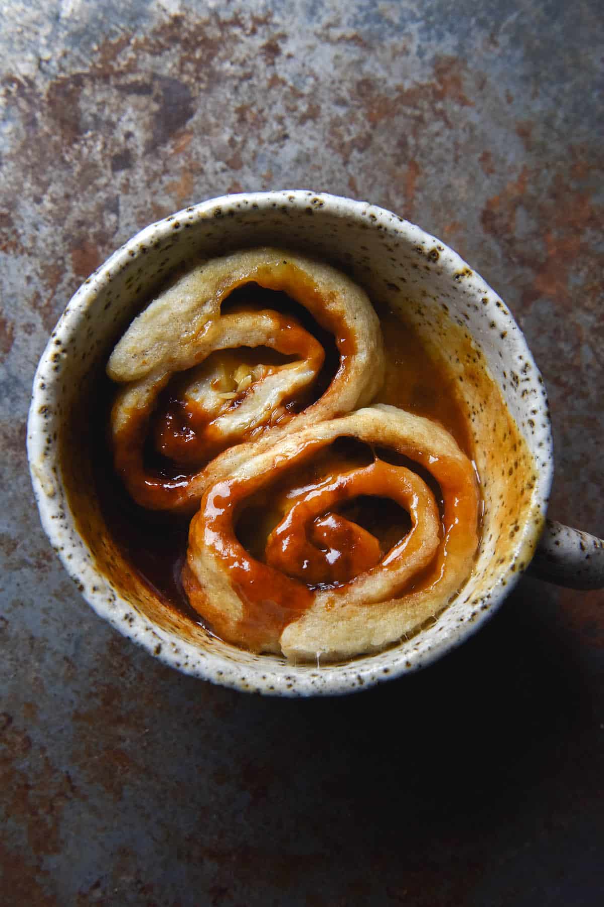 An aerial view of a gluten-free microwave Vegemite scroll in a white speckled ceramic mug. The scroll has been cut in two, revealing two Vegemite and melted cheese swirls. The cheese and Vegemite intermingle to produce varying brown, orange and yellow tones, which contrast nicely with the mottled denim blue backdrop