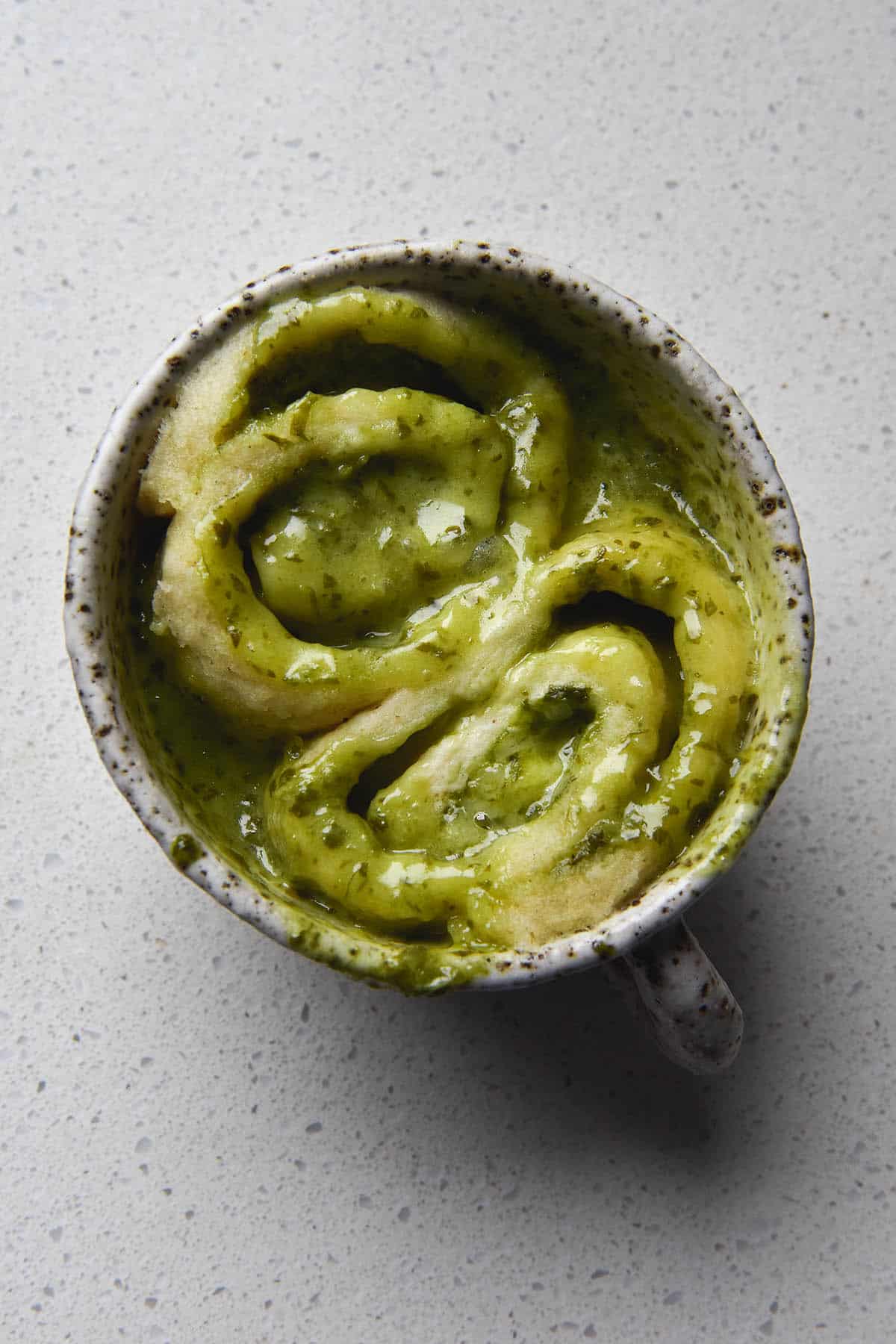 An aerial view of a gluten free vegan pesto microwave scroll in a white ceramic speckled mug atop a white kitchen countertop. The scroll has been cut in the middle before baking, revealing two cheesy pesto scrolls which sit in the mug