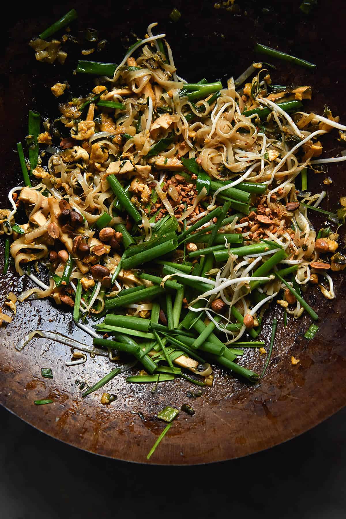 An aerial, close up view of a wok full of vegetarian Pad Thai. The noodles, tofu and egg are casually strewn about, topped with chopped spring onion chunks, garlic chives, roasted chopped peanuts and bean sprouts.