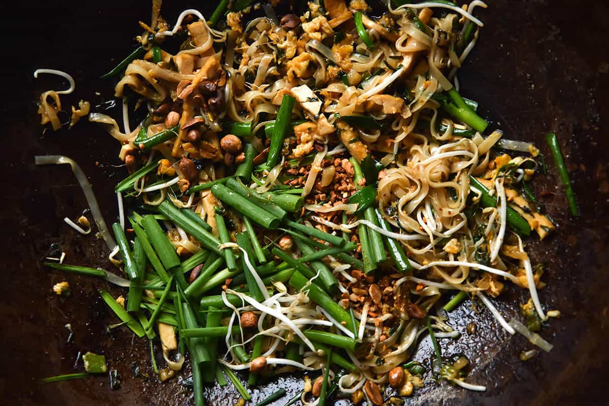 An aerial, close up view of a wok full of vegetarian Pad Thai. The noodles, tofu and egg are casually strewn about, topped with chopped spring onion chunks, garlic chives, roasted chopped peanuts and bean sprouts.