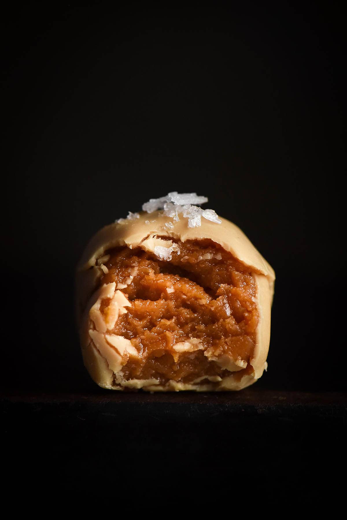 A close up side on image of a peanut butter truffle coated in white chocolate. A bite has been taken, revealing the peanut butter centre, and sea salt flakes adorn the top. The truffle sits atop a black surface against a black backdrop