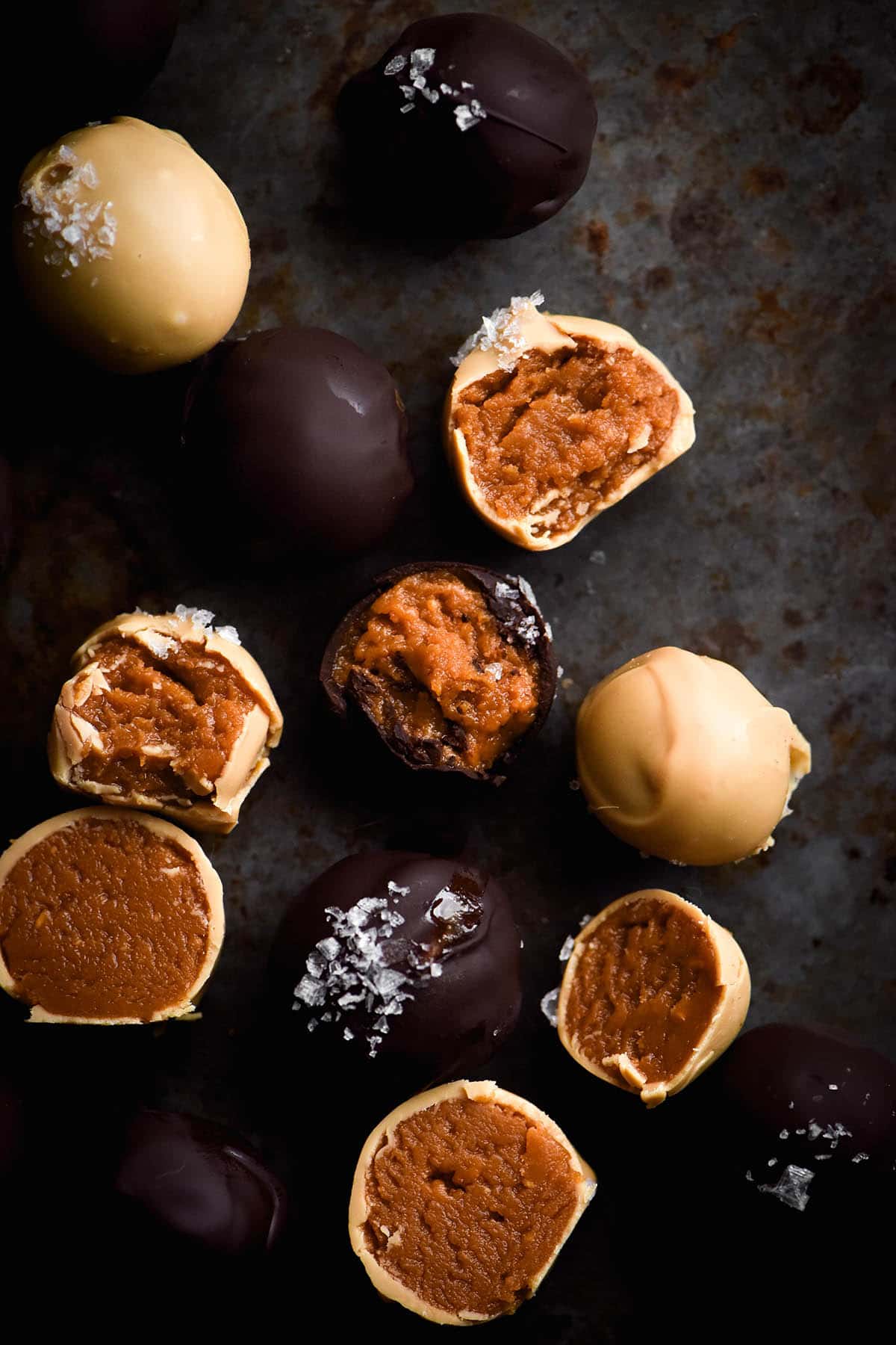 An aerial view of dark and white chocolate coated peanut truffes, some bitten into and some topped with a sprinkling of sea salt flakes. The truffles sit atop a mottled grey backdrop
