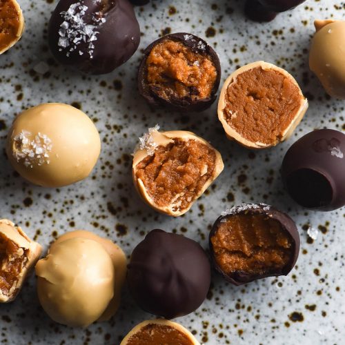 An aerial view of dark and white chocolate coated peanut truffes, some bitten into and some topped with a sprinkling of sea salt flakes. The truffles sit atop a white speckled ceramic plate