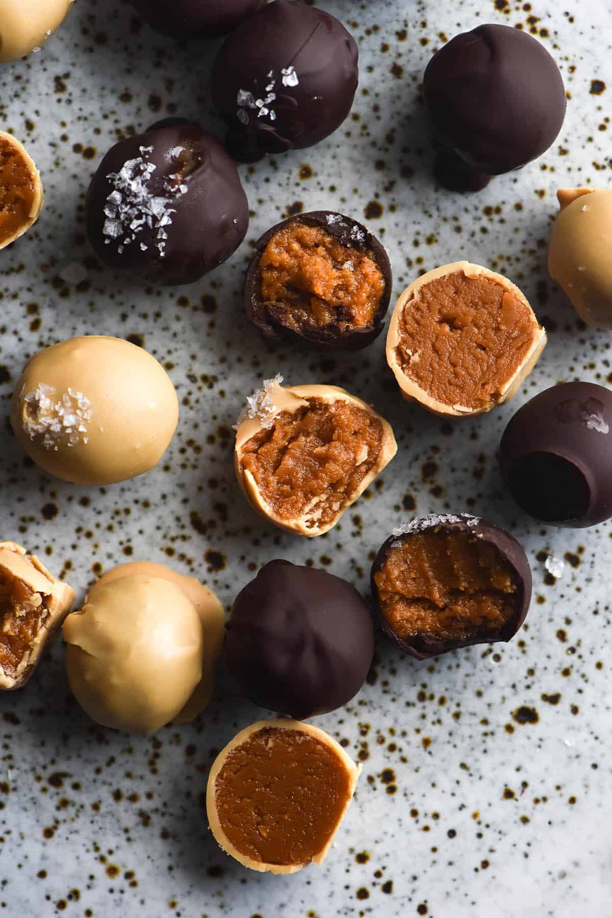An aerial view of dark and white chocolate coated peanut truffes, some bitten into and some topped with a sprinkling of sea salt flakes. The truffles sit atop a white speckled ceramic plate