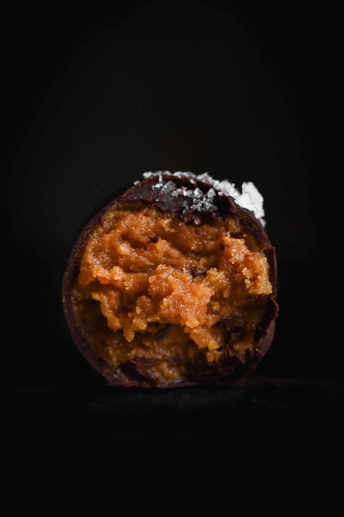 A close up side on image of a peanut butter truffle coated in dark chocolate. A bite has been taken, revealing the peanut butter centre, and sea salt flakes adorn the top. The truffle sits atop a black surface against a black backdrop