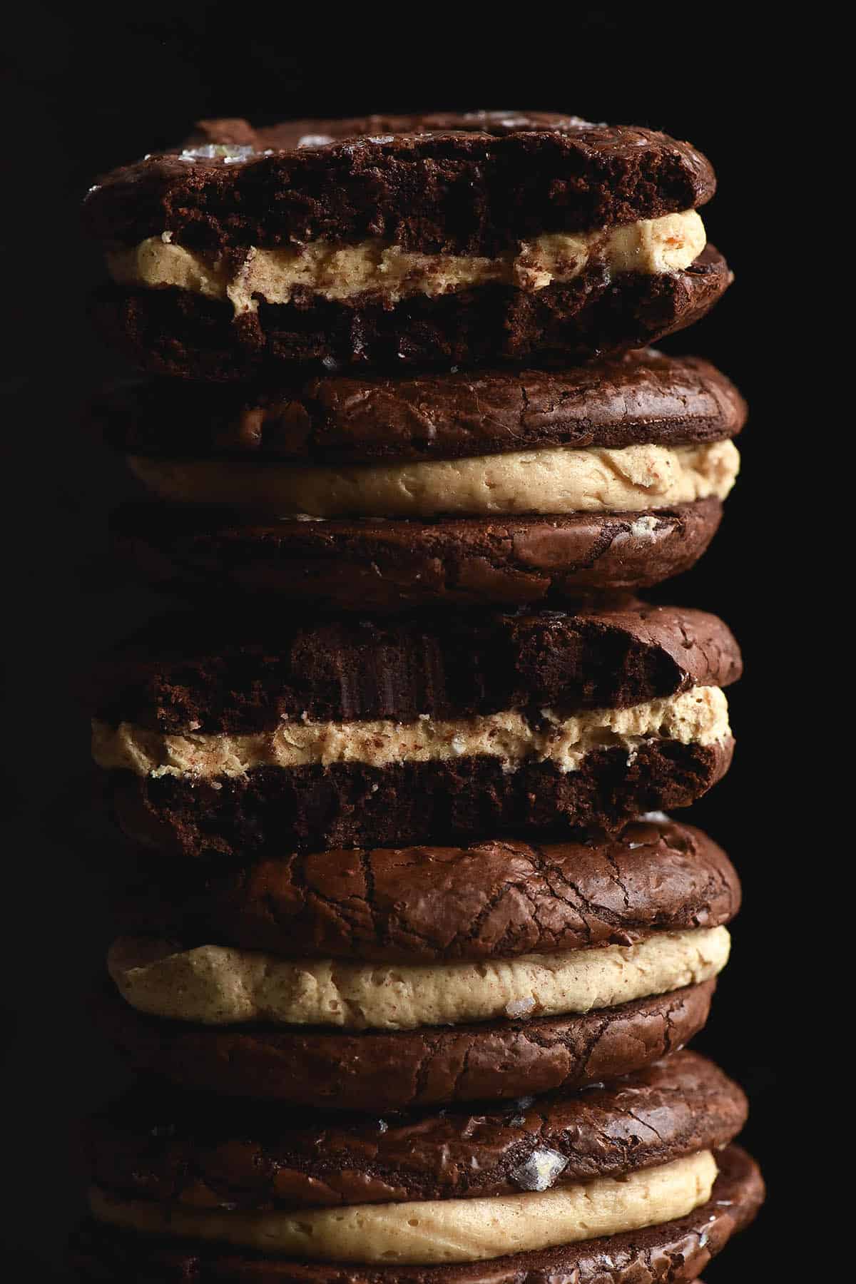 A side on, close up view of a stack of gluten free brownie cookie sandwiches. The dark background amplifies the light brown buttercream, which contrasts against the dark chocolate cookies. Specks of sea salt flakes peek out from the tops of each cookie.