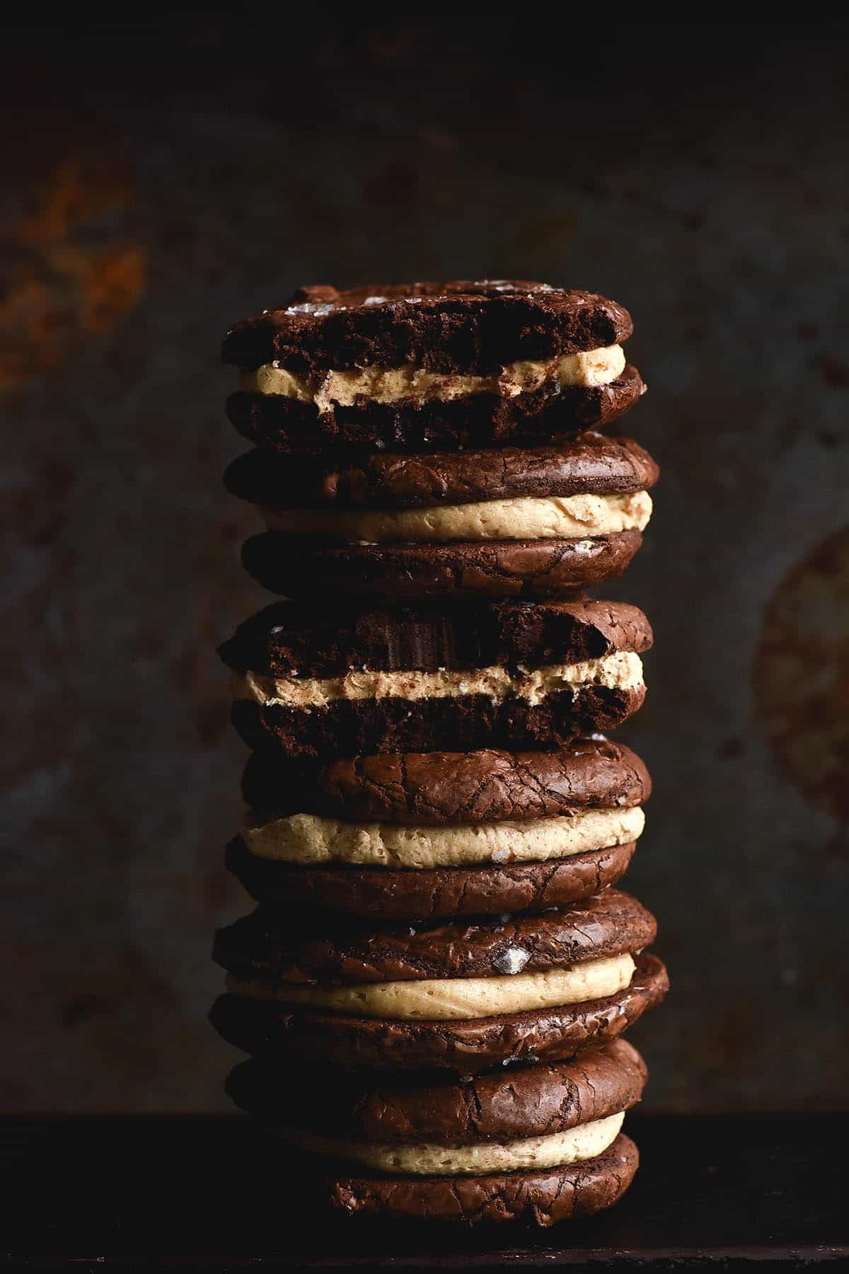 A side on moody shot of a big stack of gluten free brownie cookies made into sandwiches with a brown butter buttercream centre. The pale buttercream and a smattering of sea salt flakes contrast against the moody, rusty dark blue background