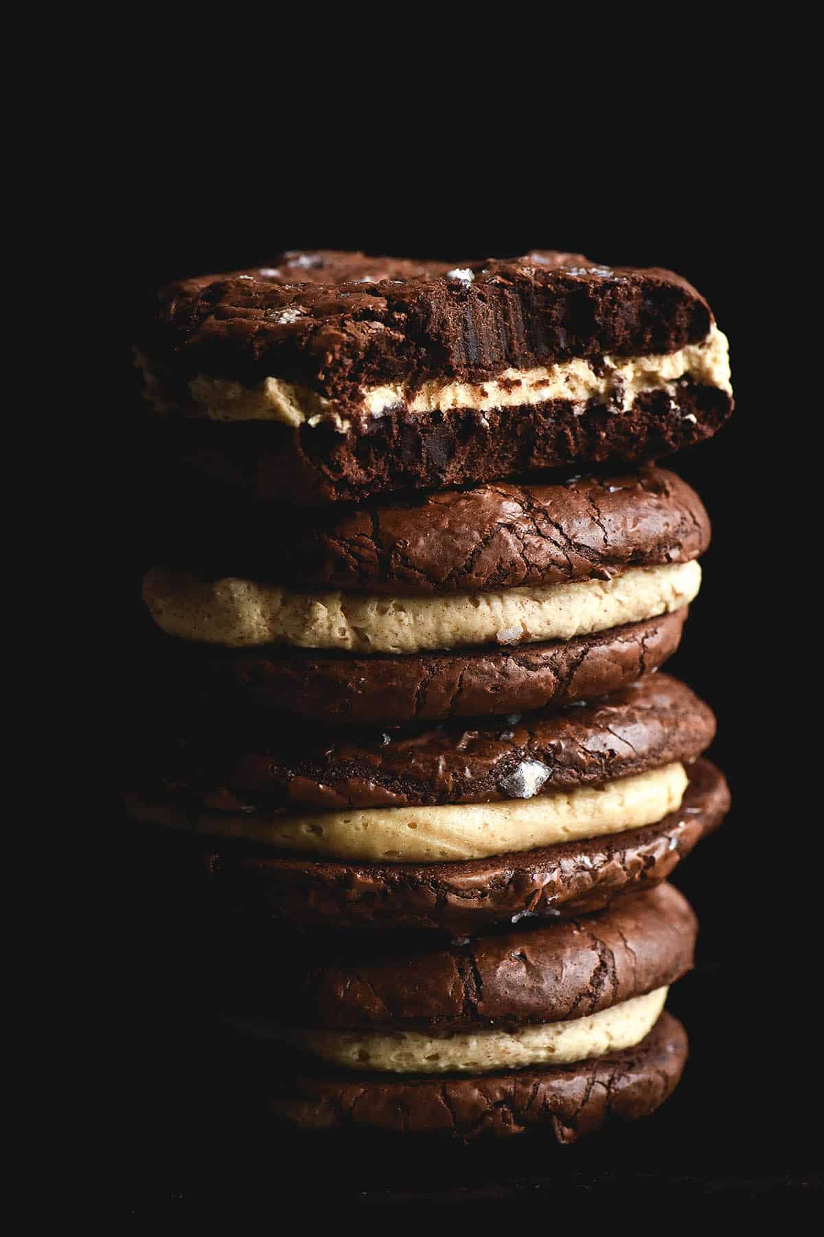 A side on moody shot of a big stack of gluten free brownie cookies made into sandwiches with a brown butter buttercream centre. The pale buttercream and a smattering of sea salt flakes contrast against the moody, rusty dark blue background