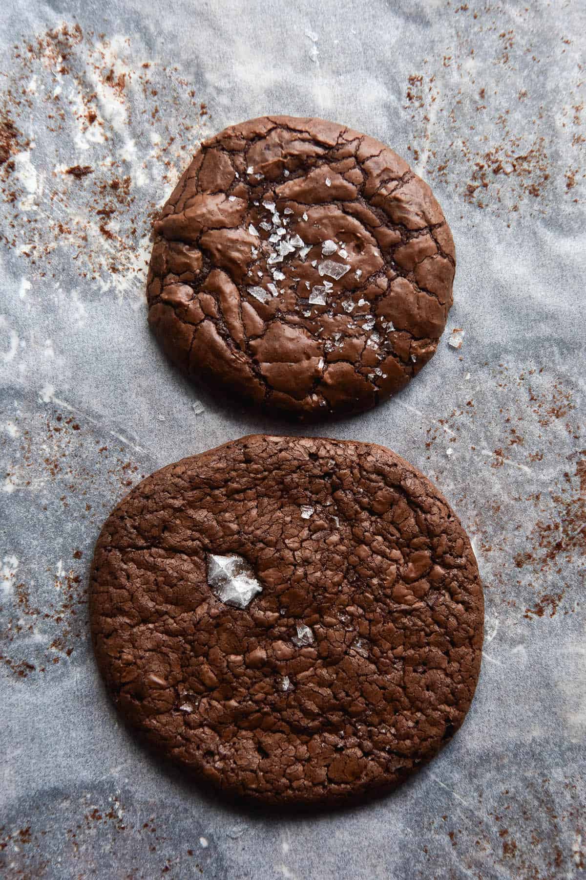 A comparative aerial image of two gluten free brownie cookies atop a baking sheet lined with parchment paper. The top cookie is shinier, cracklier and more pert, whereas the bottom cookie has a matte complexion and has spread significantly.
