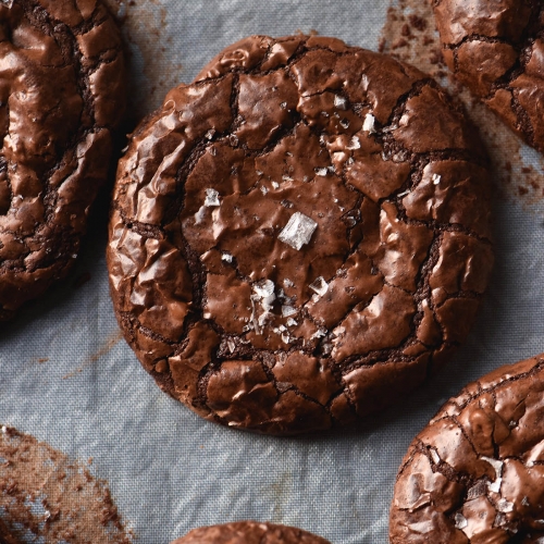 An aerial view of gluten free brownie cookies sitting atop a parchment lined baking sheet. The brownie cookies have been sprinkled with sea salt flakes that contrast against the shiny, crackled surface of the cookies.