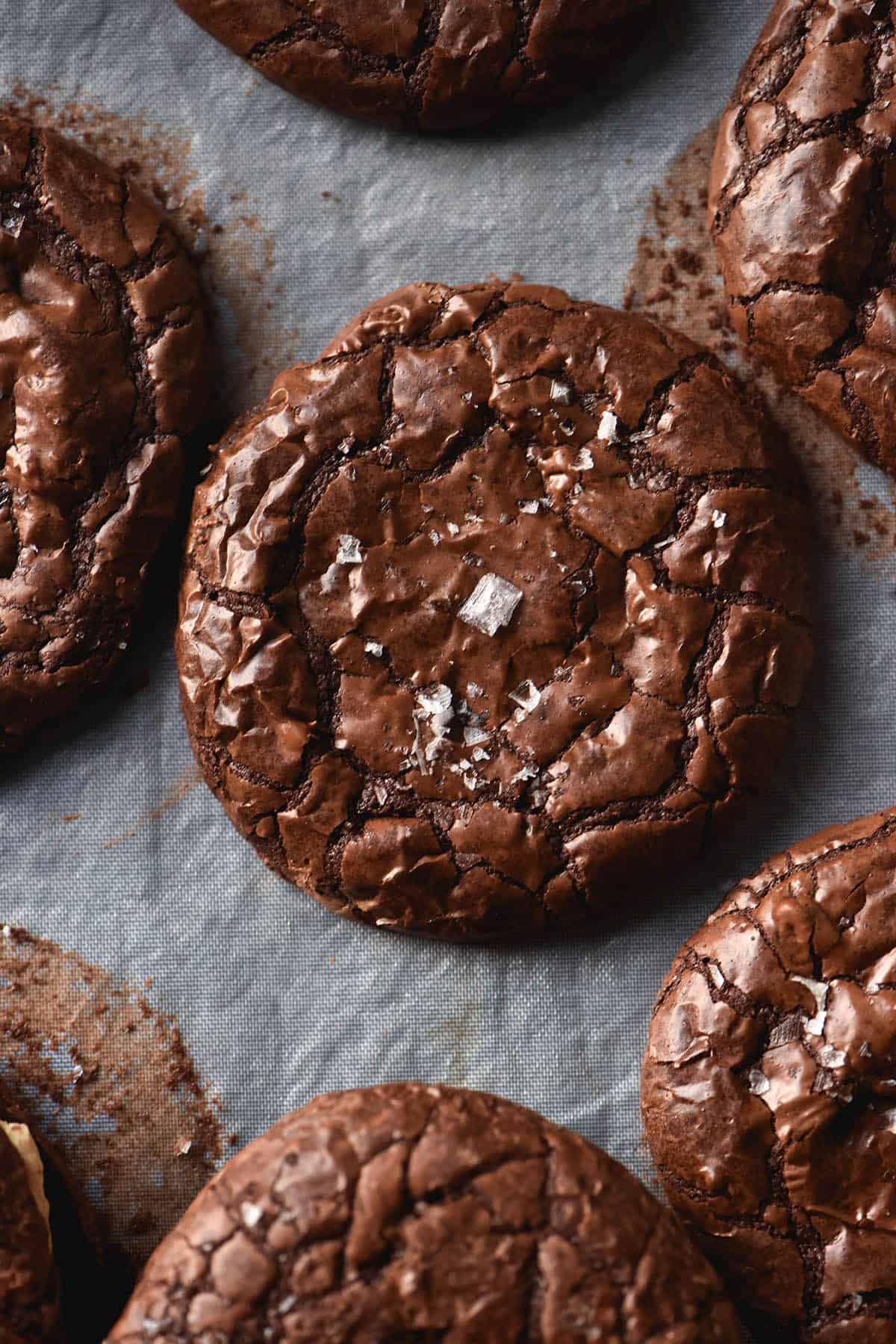 An aerial view of gluten free brownie cookies sitting atop a parchment lined baking sheet. The brownie cookies have been sprinkled with sea salt flakes that contrast against the shiny, crackled surface of the cookies.