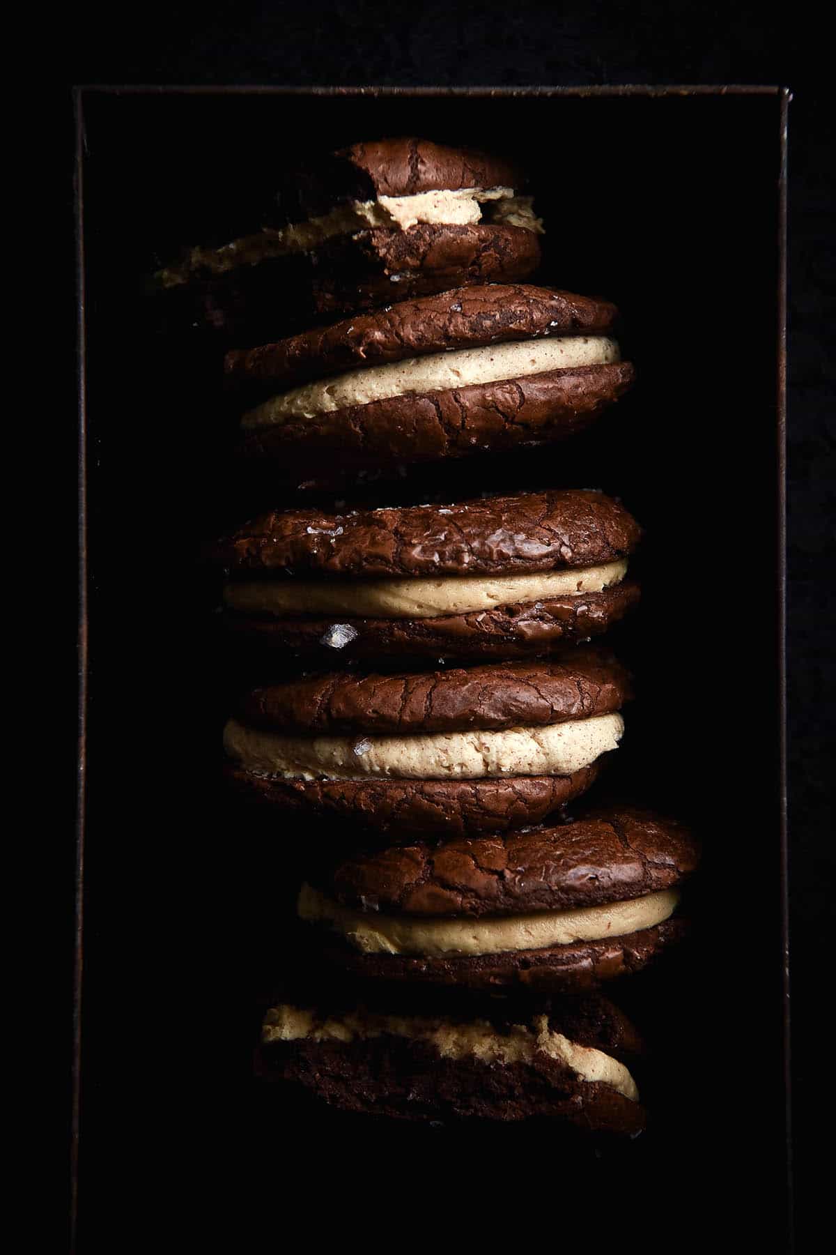 A moody aerial view of a tin of gluten free brownie sandwich cookies. The sandwich cookies are casually arranged in a vertical order, with the brown butter buttercream filling and sea salt flakes contrasting against the deep chocolate brown of the cookies