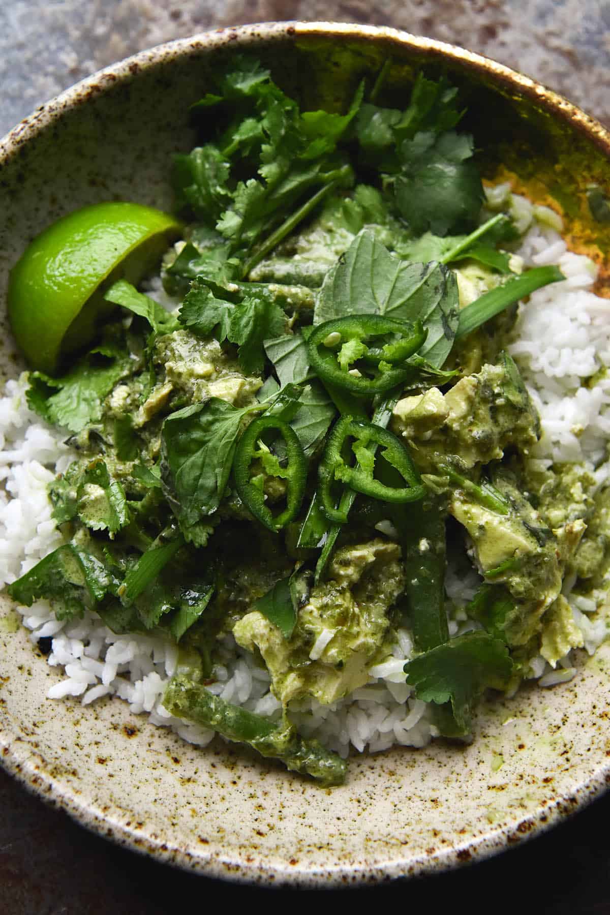 A closeup aerial image of a speckled ceramic bowl filled with white rice and topped with a FODMAP friendly, vegan Thai green curry. The curry is adorned with lots of fresh herbs and a lime wedge, and finished with three extra slices of green chilli.