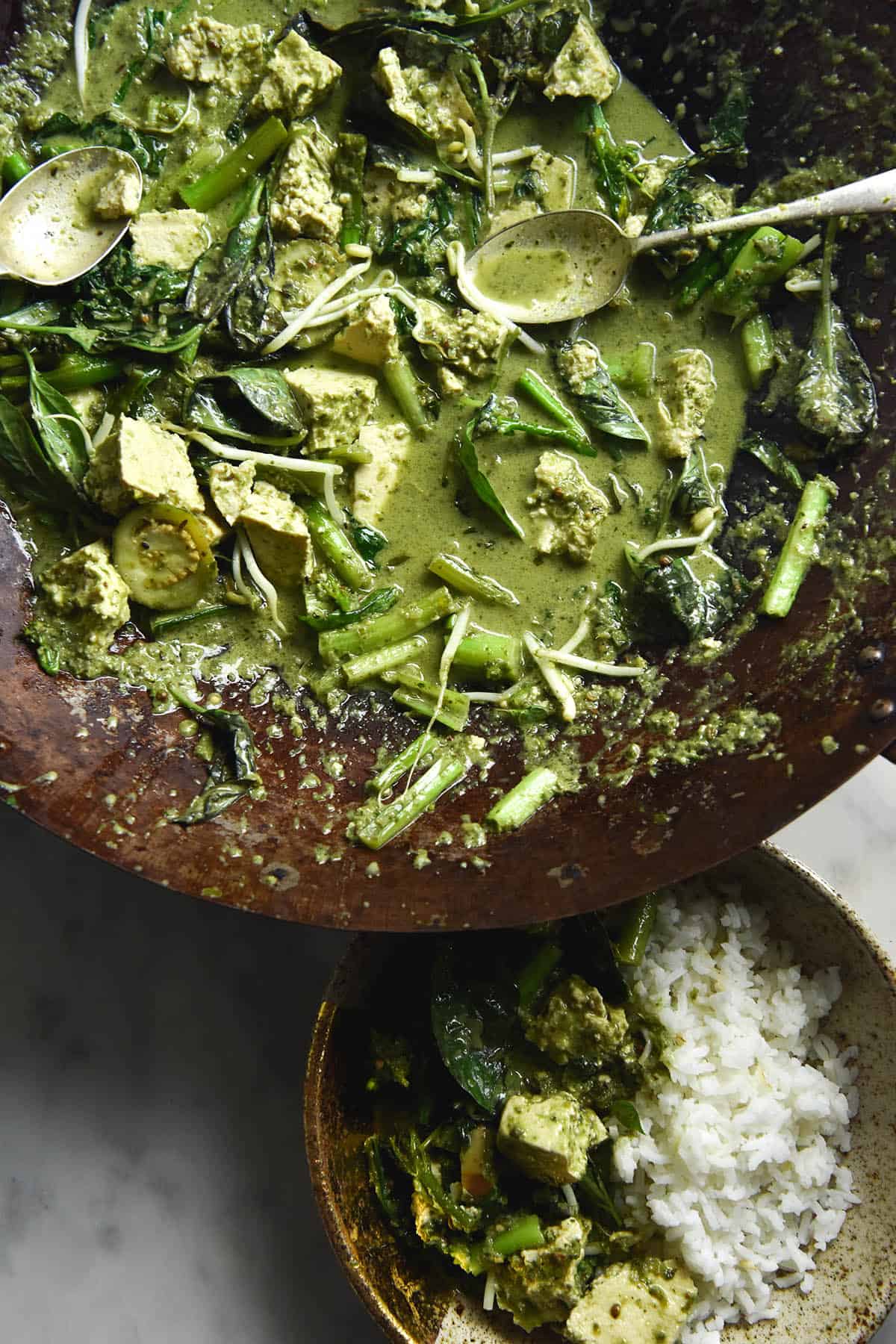 An aerial view of a wok filled with FODMAP friendly vegan Thai green curry and a plate with green curry and rice. The curry is casually strewn about the wok, with a spoon extending out to the top right hand side. It is filled with Thai eggplants, pak choi and tofu, and topped with bean sprouts and fresh herbs. The bowl sits to the bottom right of the image, filled with rice and curry, on top of a white marble table