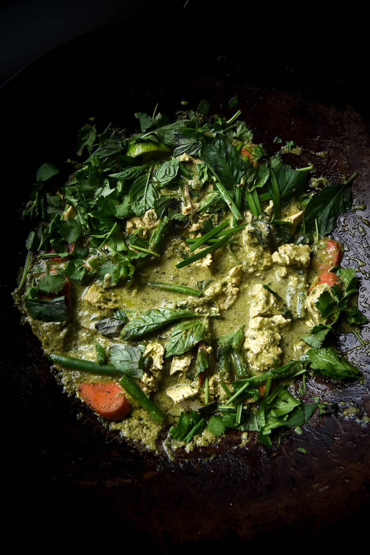 A close up aerial photo of FODMAP friendly vegan Thai curry in a wok. The curry is dotted with green beans, carrot slices and chunks of tofu, and casually adorned with lots of fresh herbs