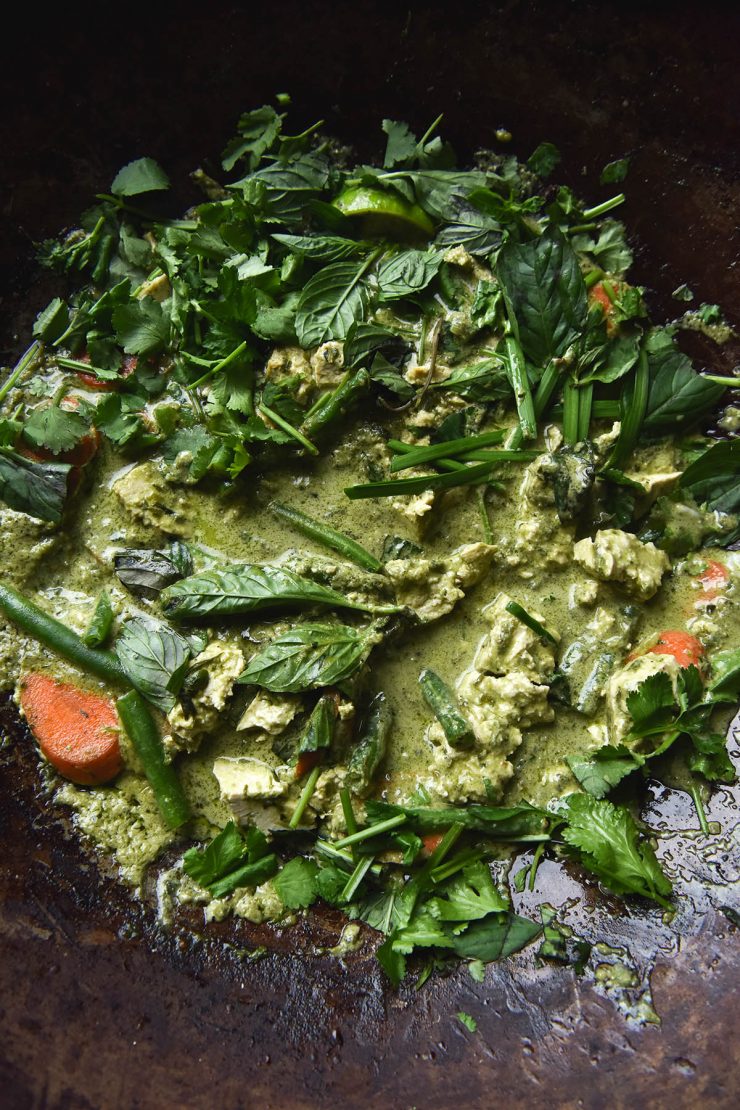 A close up aerial photo of FODMAP friendly vegan Thai curry in a wok. The curry is dotted with green beans, carrot slices and chunks of tofu, and casually adorned with lots of fresh herbs