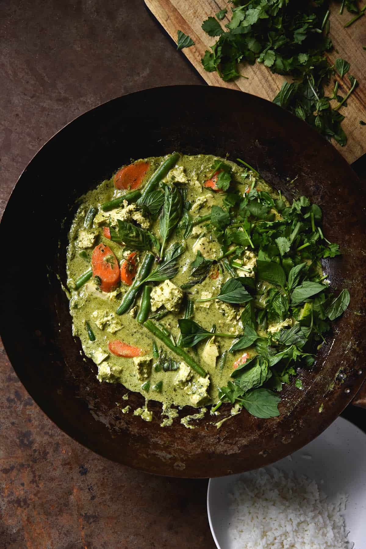 An aerial view of a wok filled with FODMAP friendly vegan Thai green curry. The curry is adorned with lots of fresh herbs, which also sit on a chopping board in the top right hand of the image. A bowl of rice sits in the bottom right hand. The scene is set on a rusty coloured backdrop