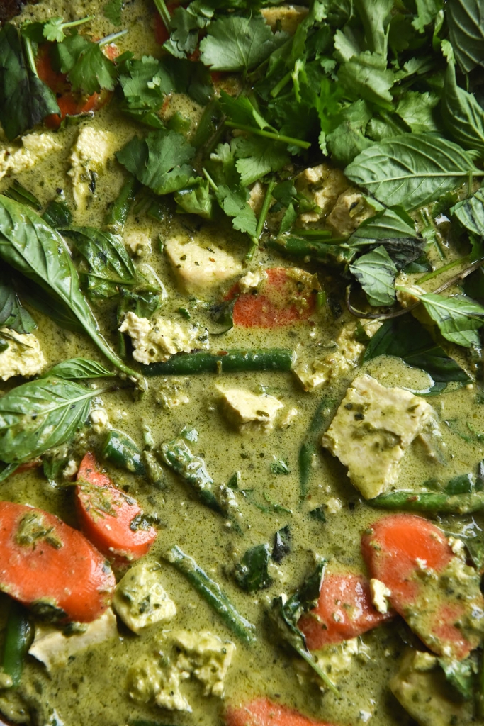 A close up aerial view of FODMAP friendly Thai green curry. The curry is a verdant green and dotted with sliced carrot, chunks of tofu and green beans. A pile of fresh herbs sits in the top left of the image atop the curry