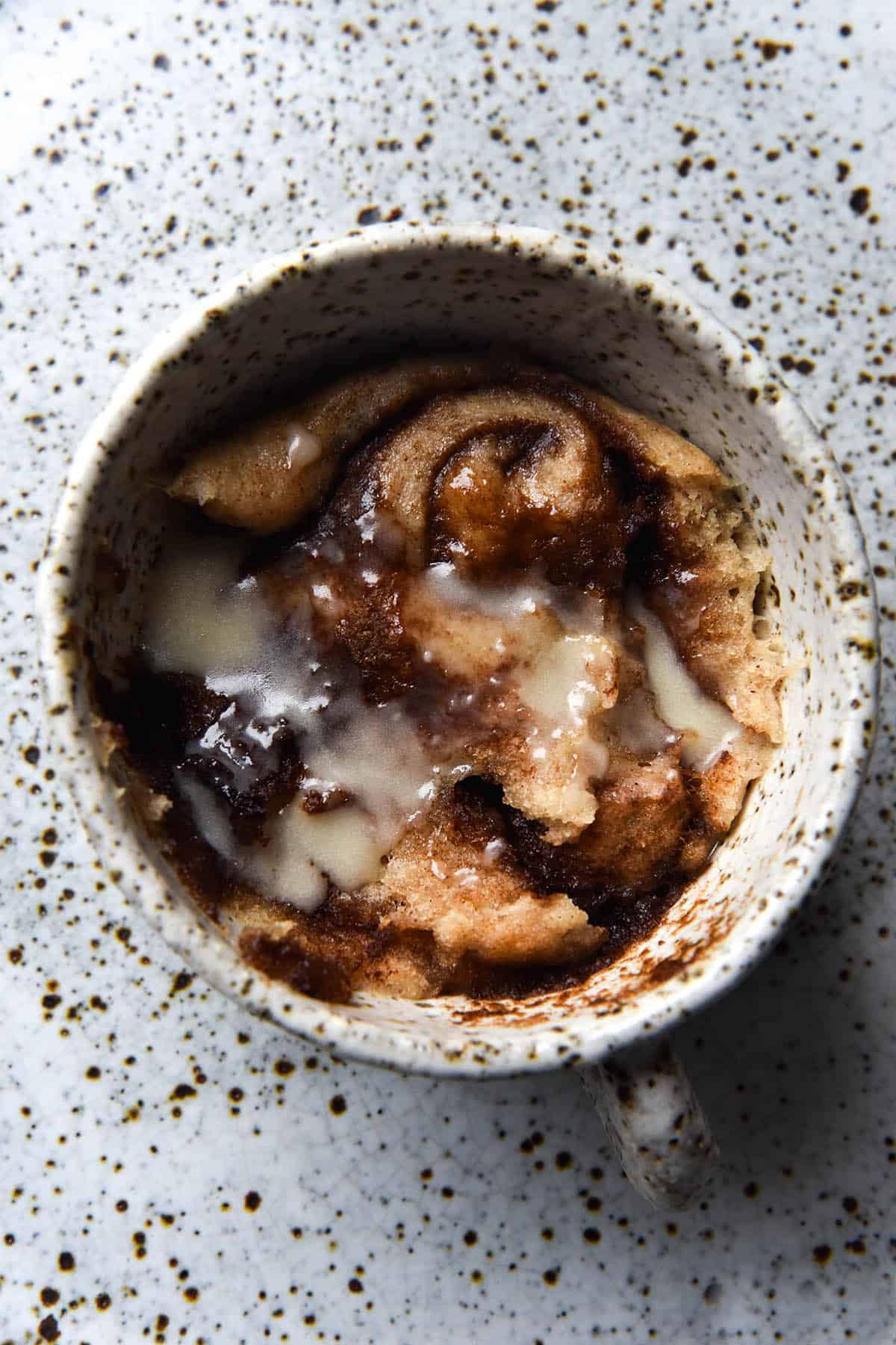 An aerial close up view of a gluten-free vegan microwave cinnamon scroll. The scroll has been cooked in a white speckled ceramic mug, which sits atop a white speckled ceramic plate. The scroll has been eaten into and swirls of cinnamon are revealed underneath the dough. It has been drizzled with an icing sugar glaze