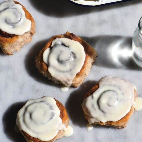 An aerial view of gluten-free vegan cinnamon scrolls strewn casually across a white marble table. The scrolls are iced and sit in harsh sunlight, surrounded by two water glasses which cast their shadow and light over the image. A tray of more un-iced scrolls sit to the top right of the image