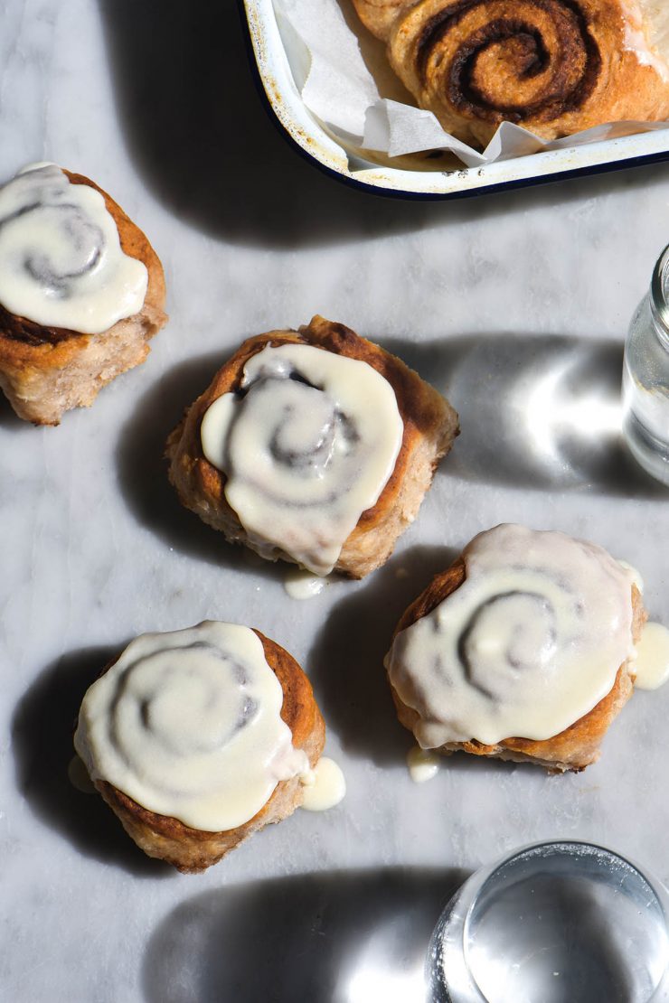 An aerial view of gluten-free vegan cinnamon scrolls strewn casually across a white marble table. The scrolls are iced and sit in harsh sunlight, surrounded by two water glasses which cast their shadow and light over the image. A tray of more un-iced scrolls sit to the top right of the image
