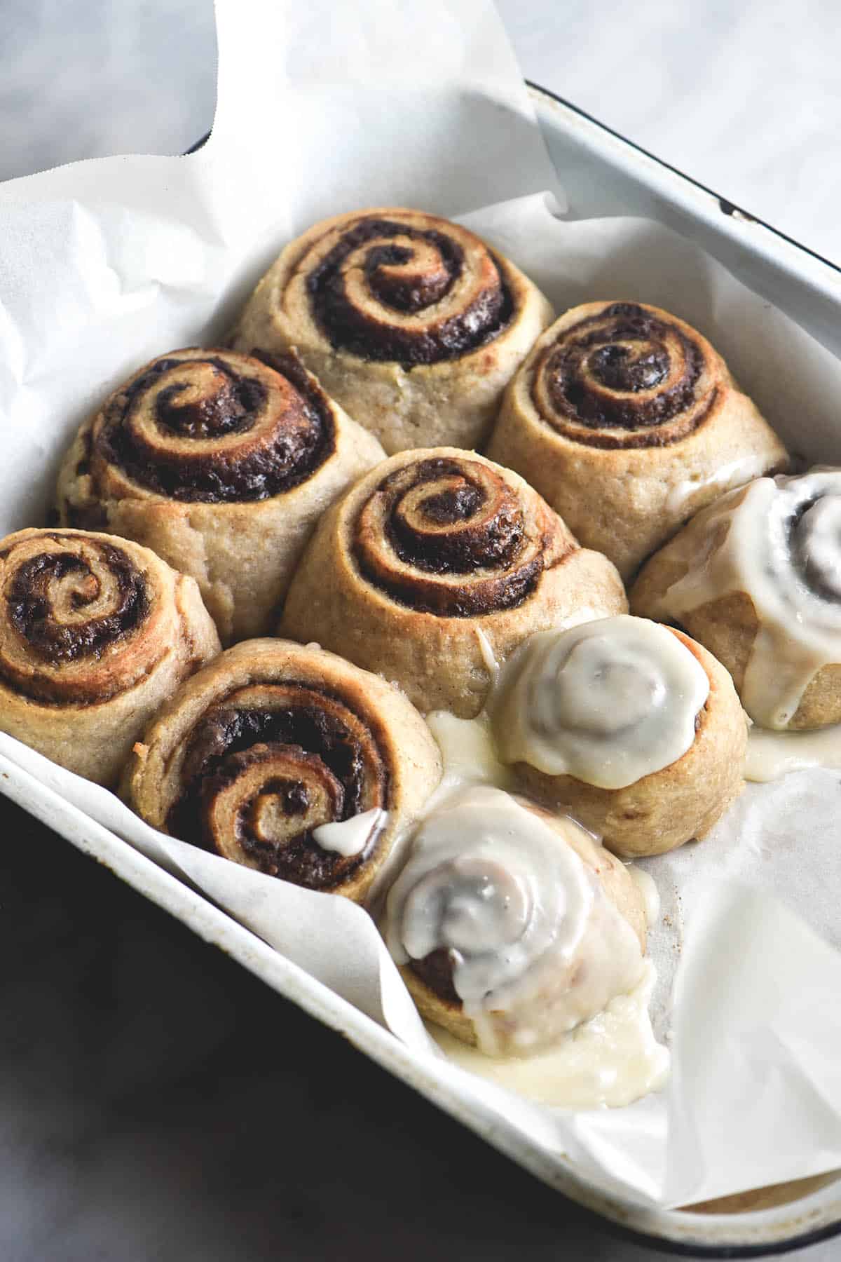 A tray of vegan, gluten free cinnamon scrolls sitting on a white marble table. The row of scrolls to the right of the image have been casually iced, while the two rolls of scrolls tp the left of the image are bare, exposing their cinnamon swirls