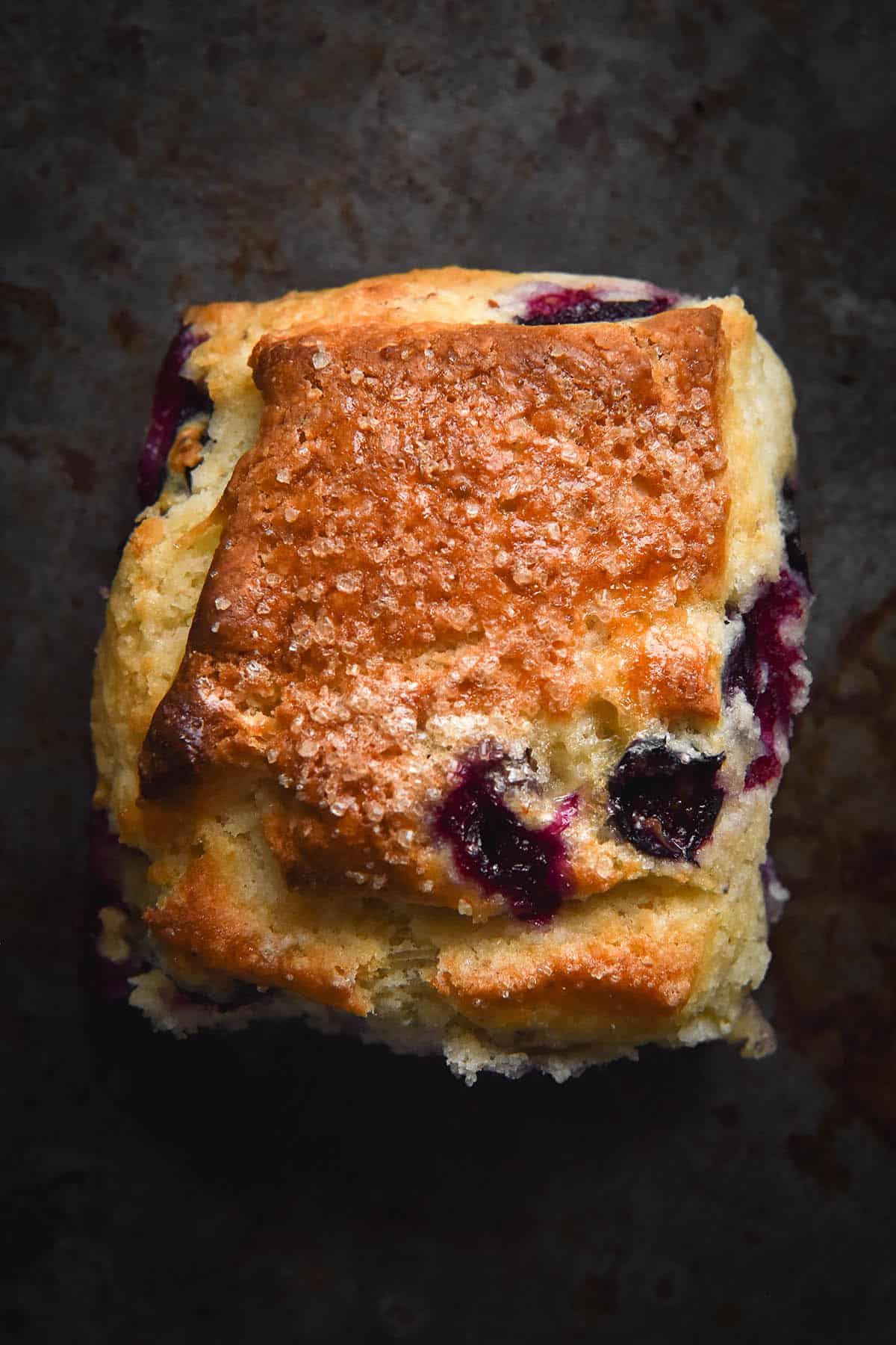 An aerial close up view of a gluten free blueberry scones against a dark metal backdrop. The scone is a square shape and has oozy blueberries dotted across the golden top, which is also sprinkled with finishing sugar