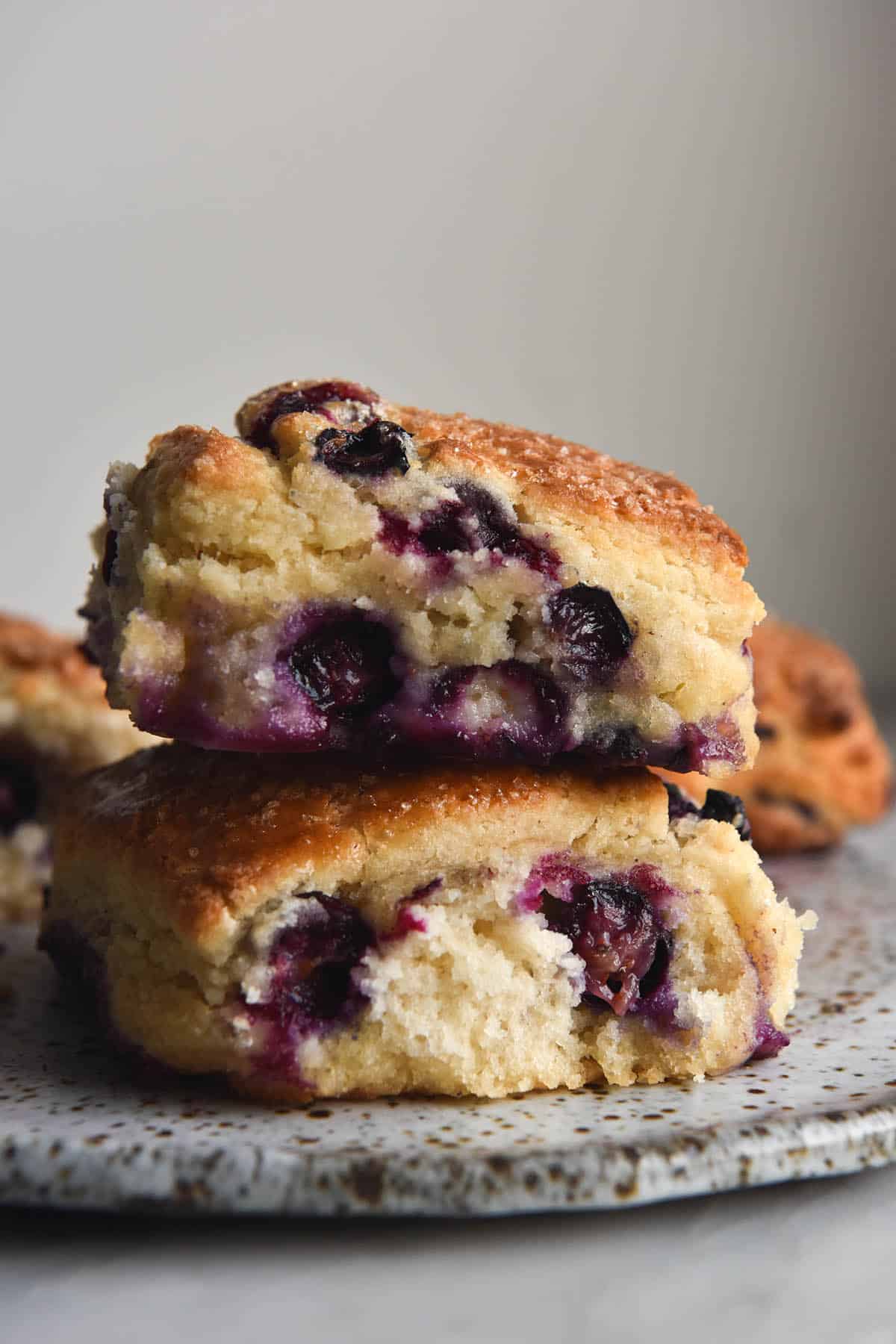 An side on view of a stack of two gluten free blueberry scones. They sit atop a white speckled ceramic plate against a white wall. The scones are fluffy and dotted with oozy blueberries. Scones sit to both the left and right of the stack in the background