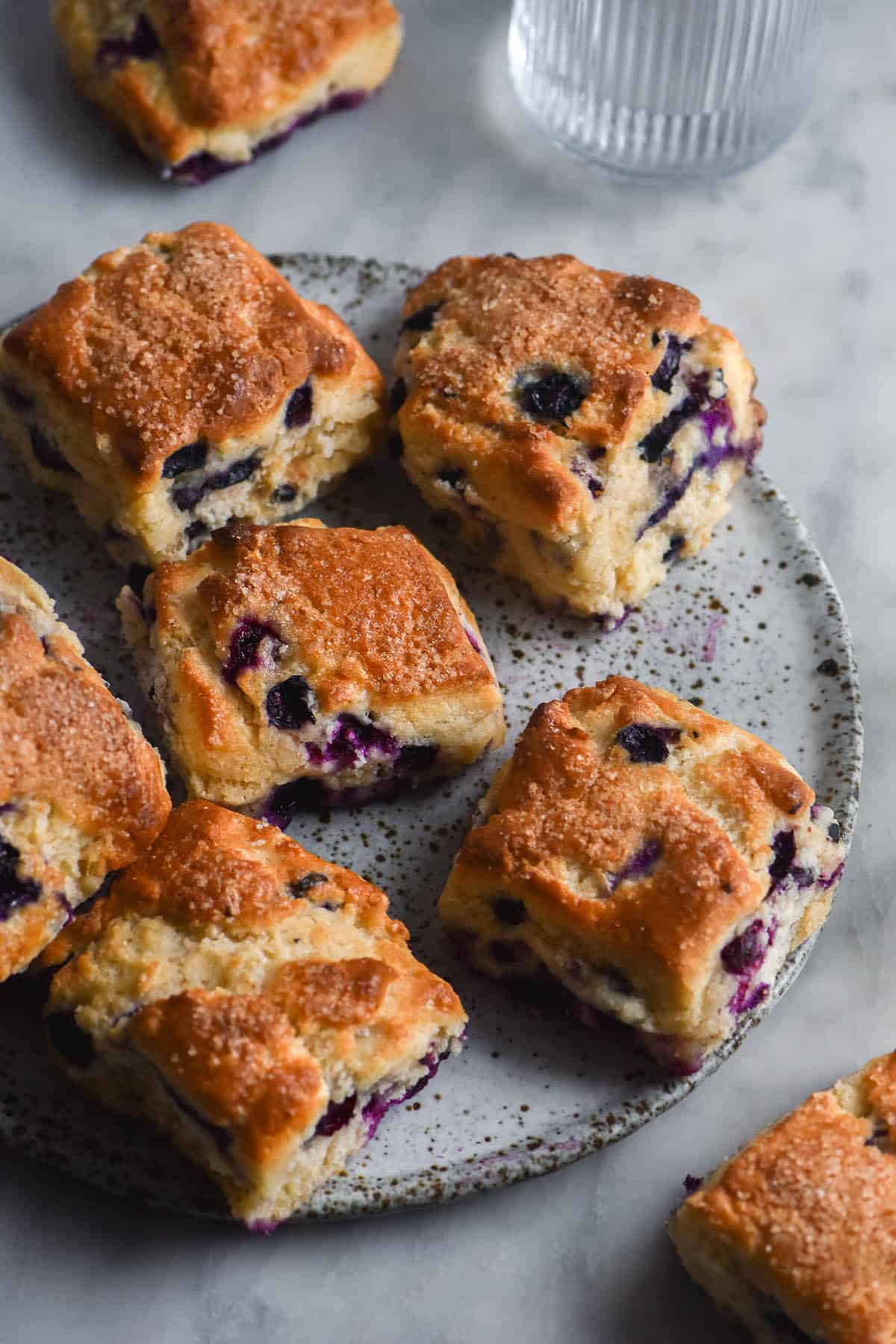 An aerial, side on view of a white marble table strewn with gluten free blueberry scones. A white speckled ceramic plate sits in the middle of the table and is topped with scones, but scones also sit on the table surrounding the plate. A half filled water glass sits to the top right hand side of the image.