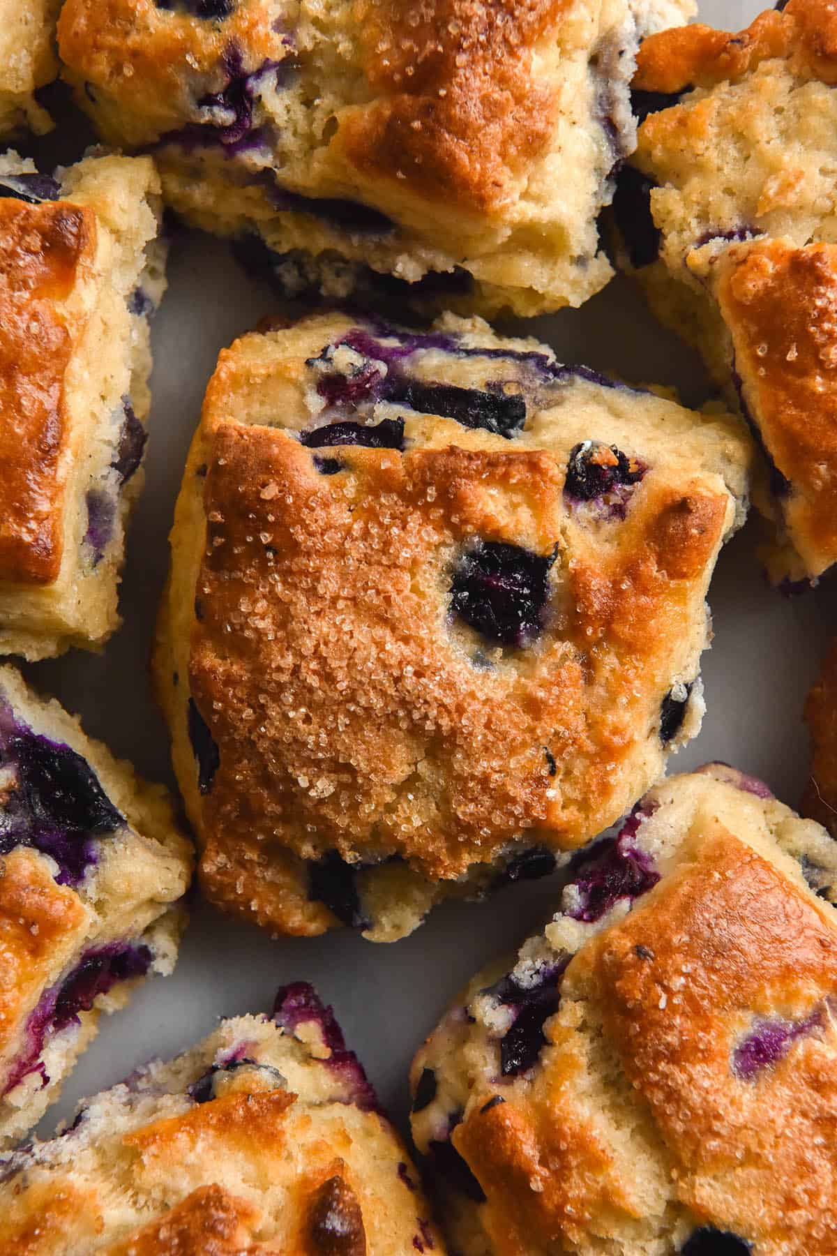 An aerial, close up shot of gluten free blueberry scones. The scones are golden brown on top, with finishing sugar and oozy blueberries stud the tops.